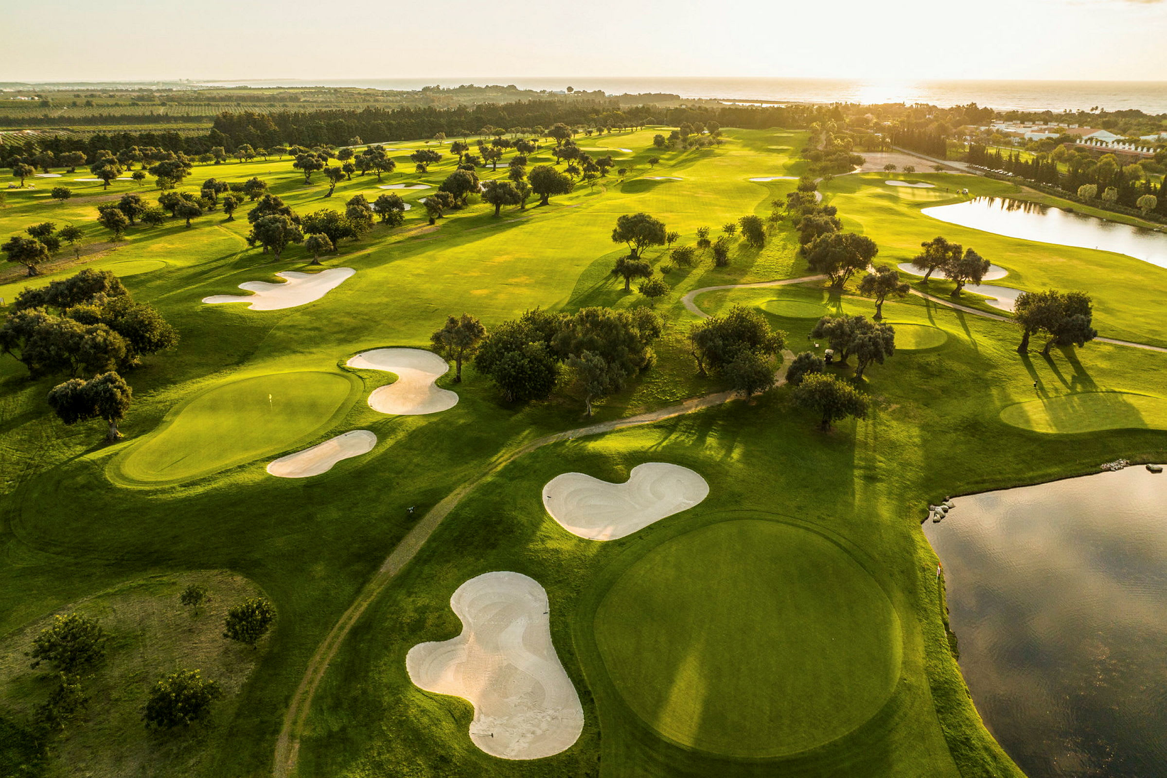 Flyutsikt over en golfbane med grønne fairways, sandbunkere og vannhindringer ved solnedgang. (AI-generert tekst)