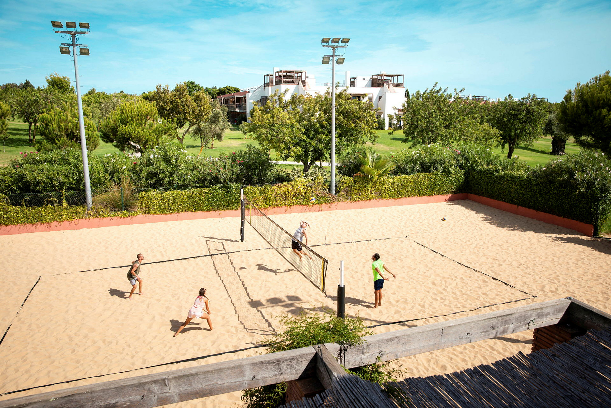 Sandvolleyballbane med fire personer som spiller, omgitt av grønt landskap og bygninger i bakgrunnen. (AI-generert tekst)