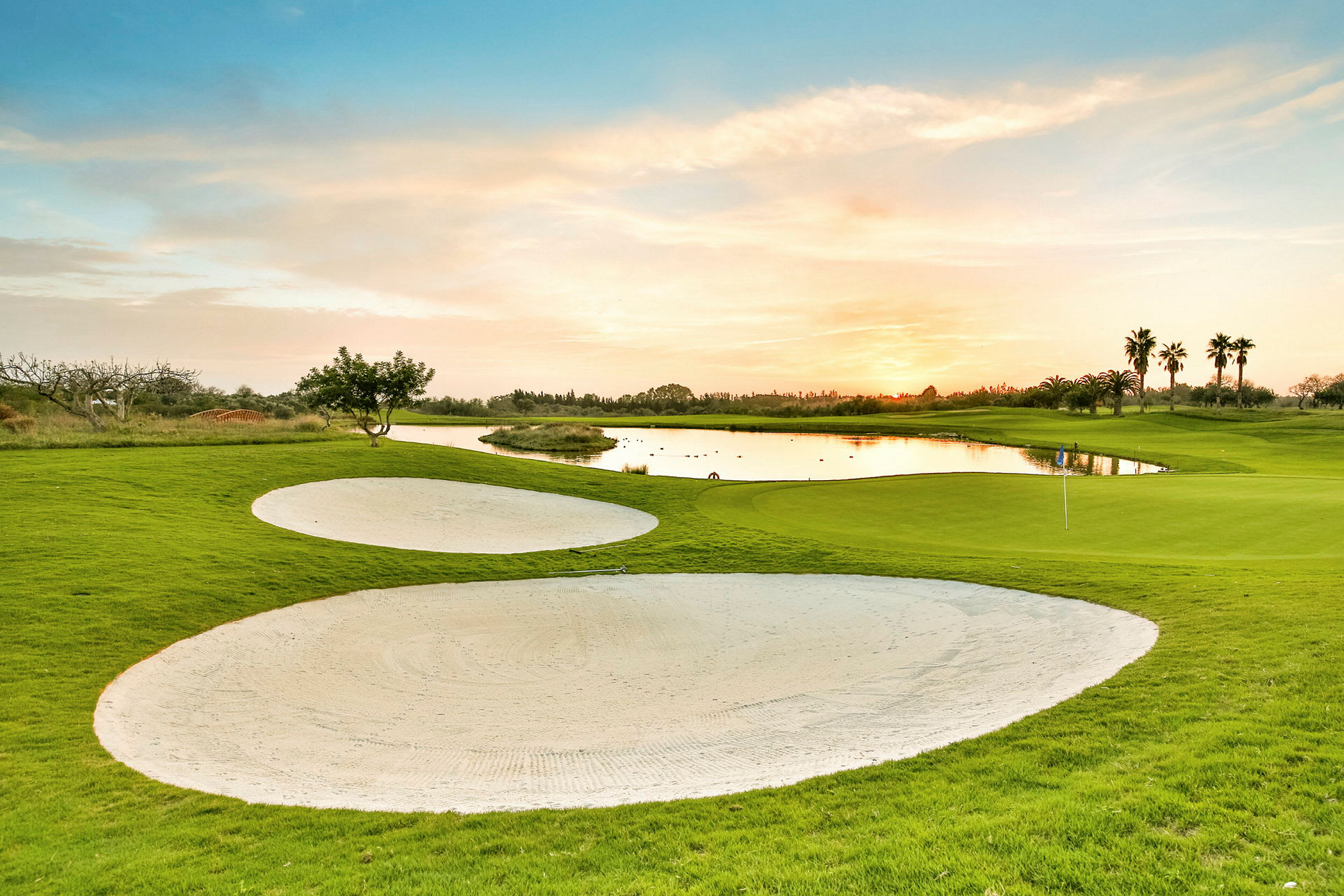 En golfbane med grønt gress, sandbunkere og en dam ved solnedgang. (AI-generert tekst)