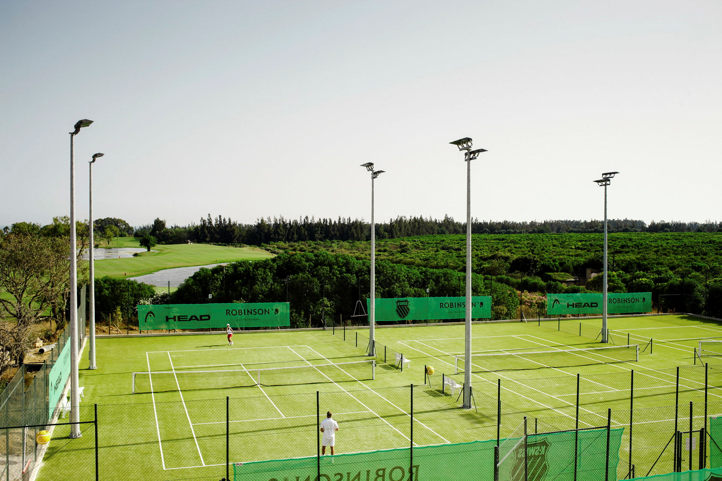 Tennisbaner med grønt underlag omgitt av landskap. (AI-generert tekst)