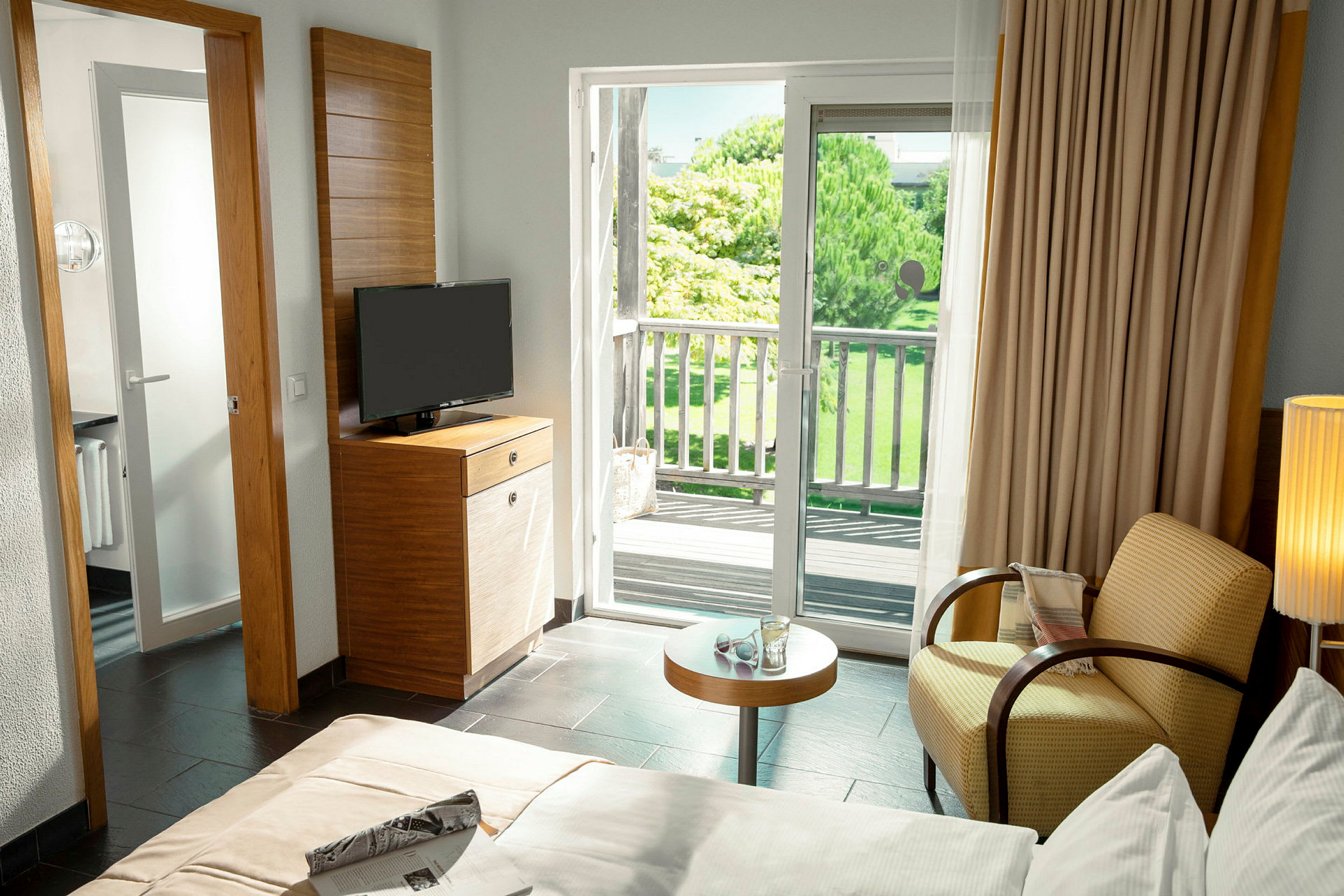Et hotellrom med seng, lenestol, bord, TV og en balkong som vender mot en frodig hage. (AI-generert tekst)