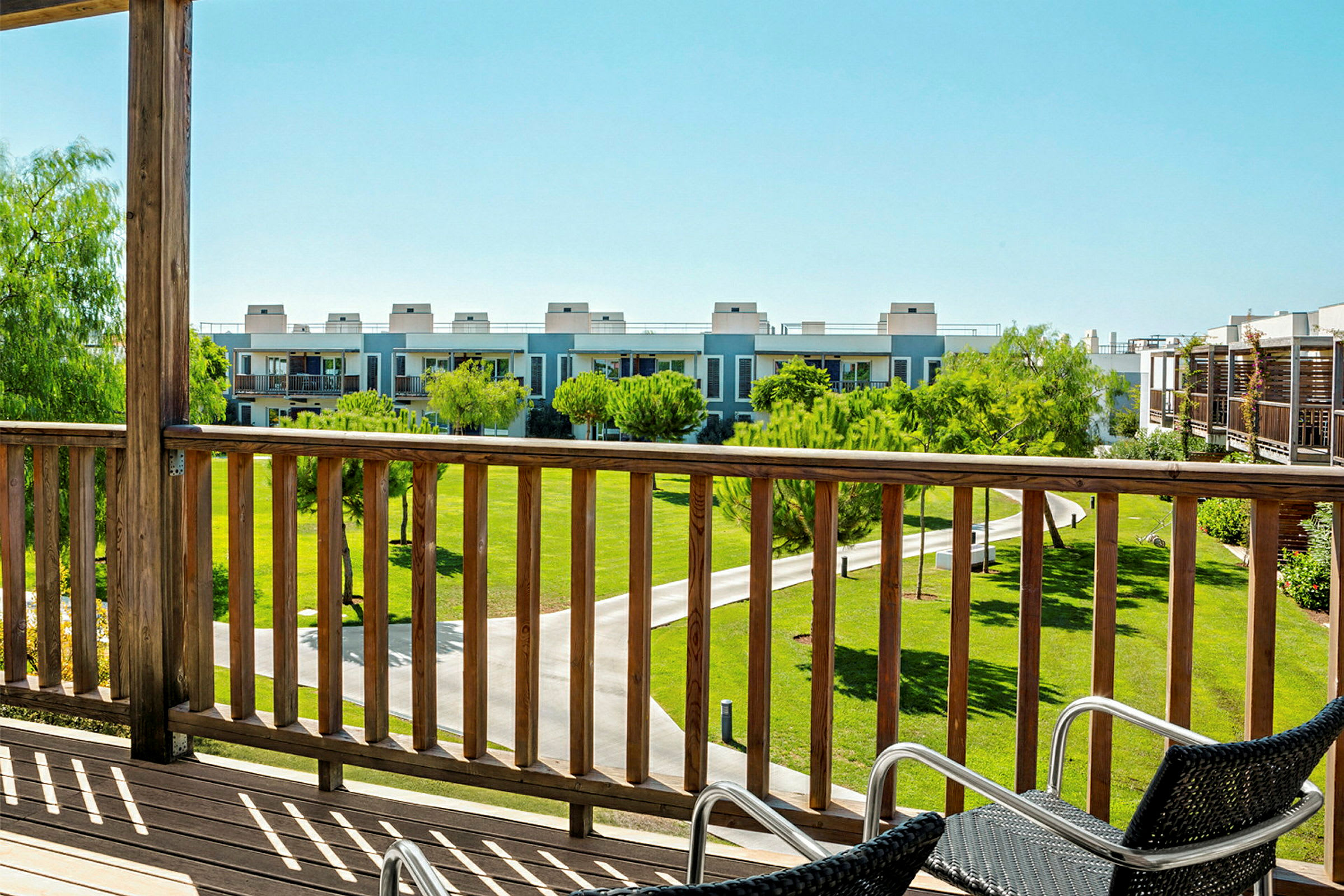 Utsikt fra en terrasse med trerekkverk over en grønn plen og moderne bygninger i bakgrunnen. (AI-generert tekst)