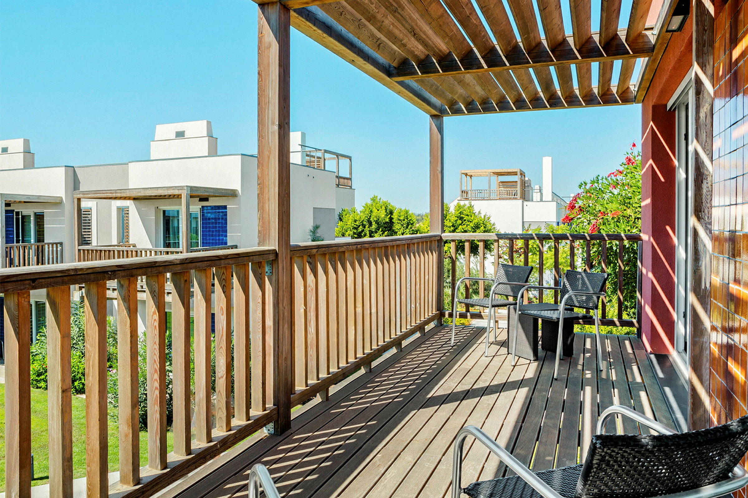 En balkong med trerekkverk og pergola med utsikt over andre bygninger og grøntområder. Stoler og et lite bord er på balkongen. (AI-generert tekst)