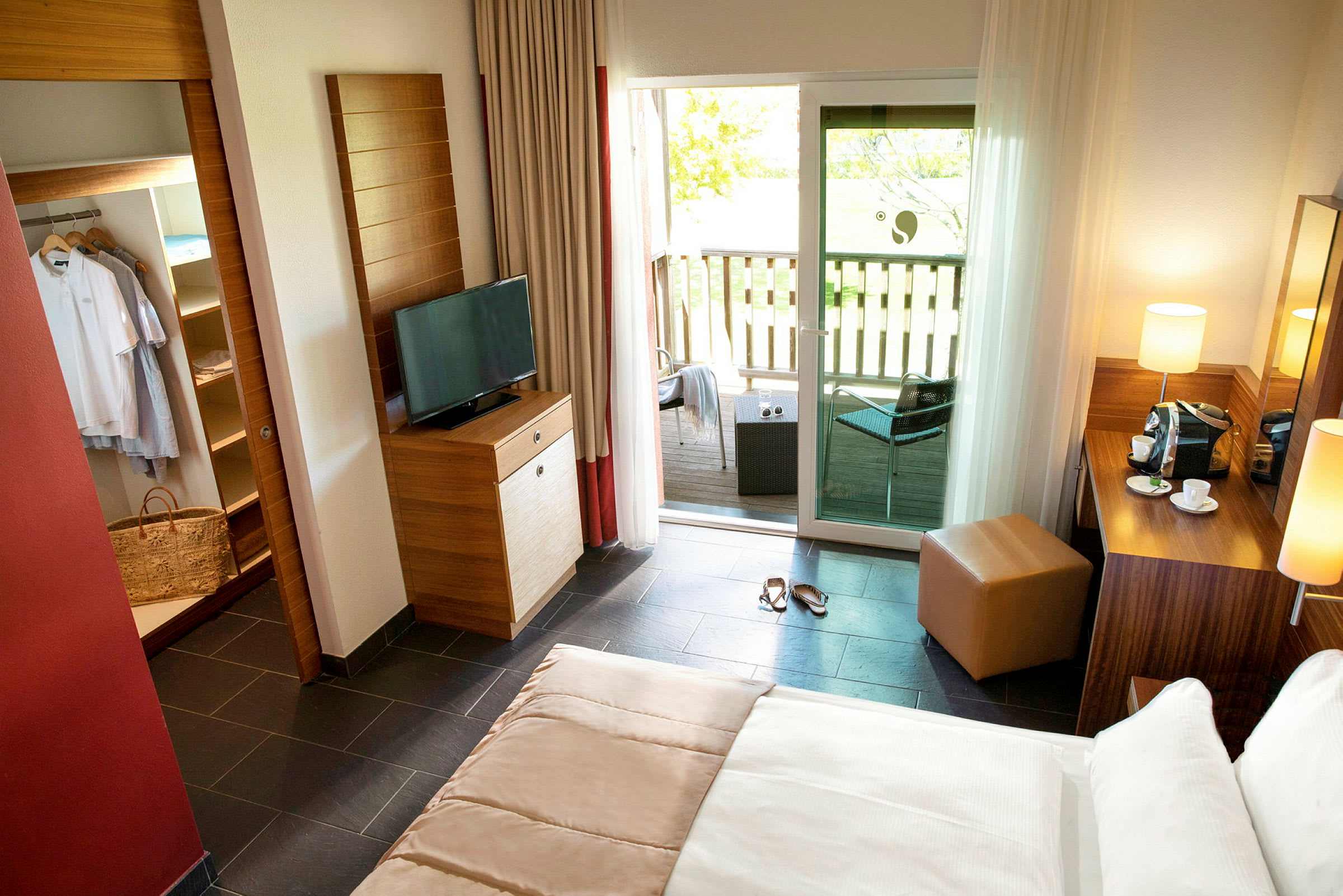Et lyst hotellrom med seng, TV, en garderobe og en balkong med sittegruppe utenfor. (AI-generert tekst)