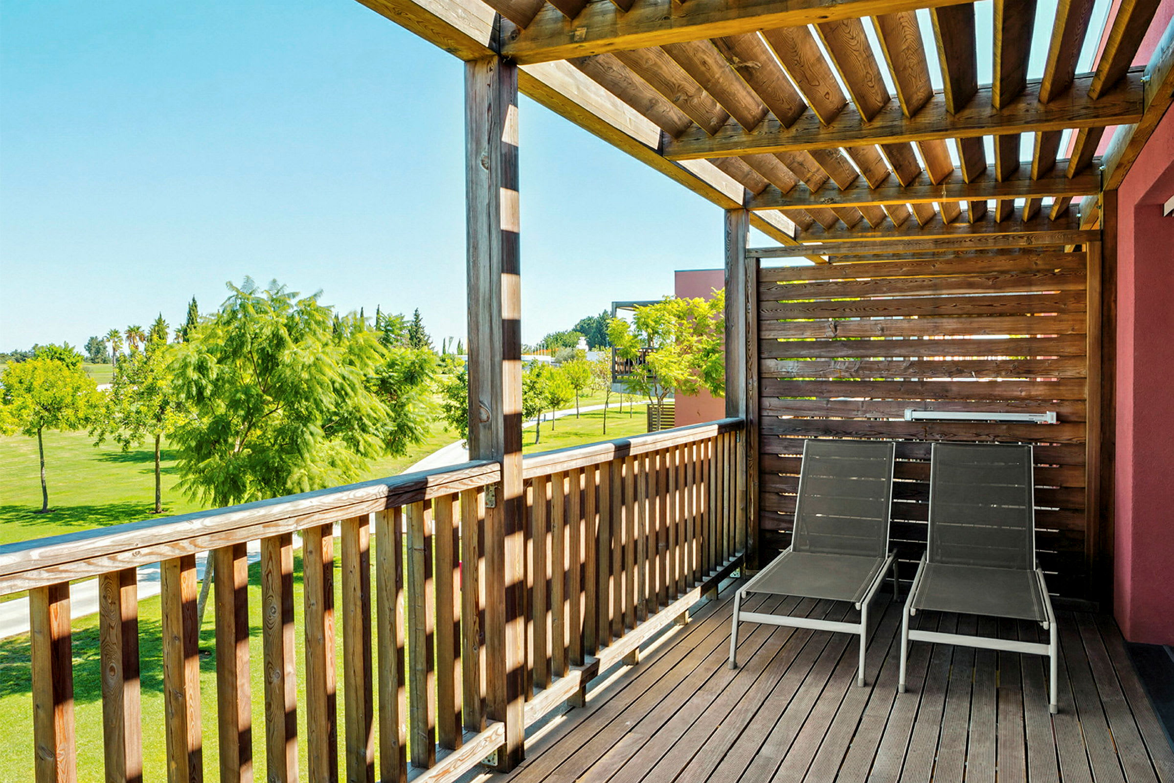 En treterrasse med to solsenger plassert under et tak av trelameller, omkranset av et inngjerding, med utsikt over et grønt felt og noen trær. (AI-generert tekst)