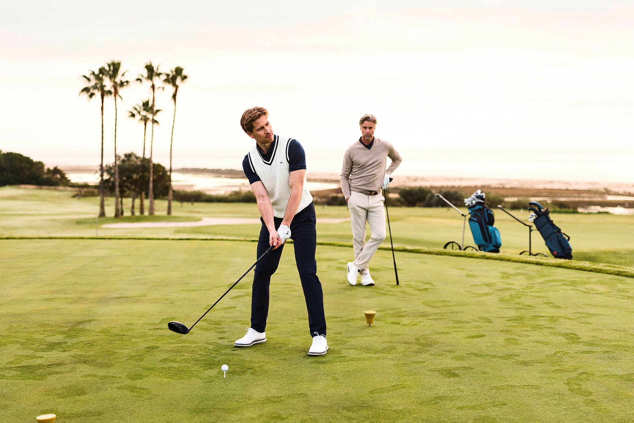 En person spiller golf og en annen står ved siden av, med en grønn golfbane i bakgrunnen. (AI-generert tekst)