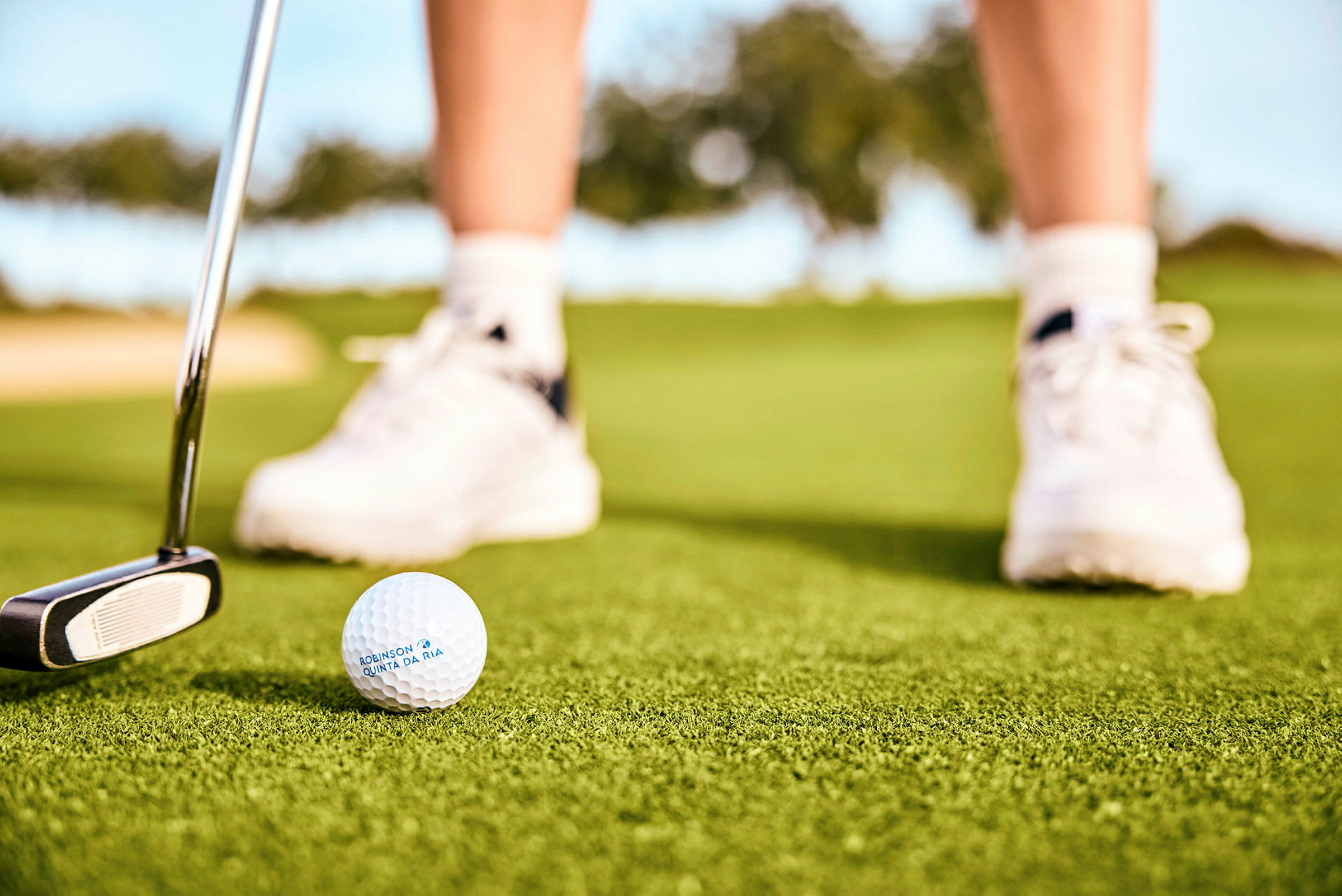 En golfball og en putter på en grønn gressplen, med en persons føtter iført hvite sportssko i bakgrunnen. (AI-generert tekst)