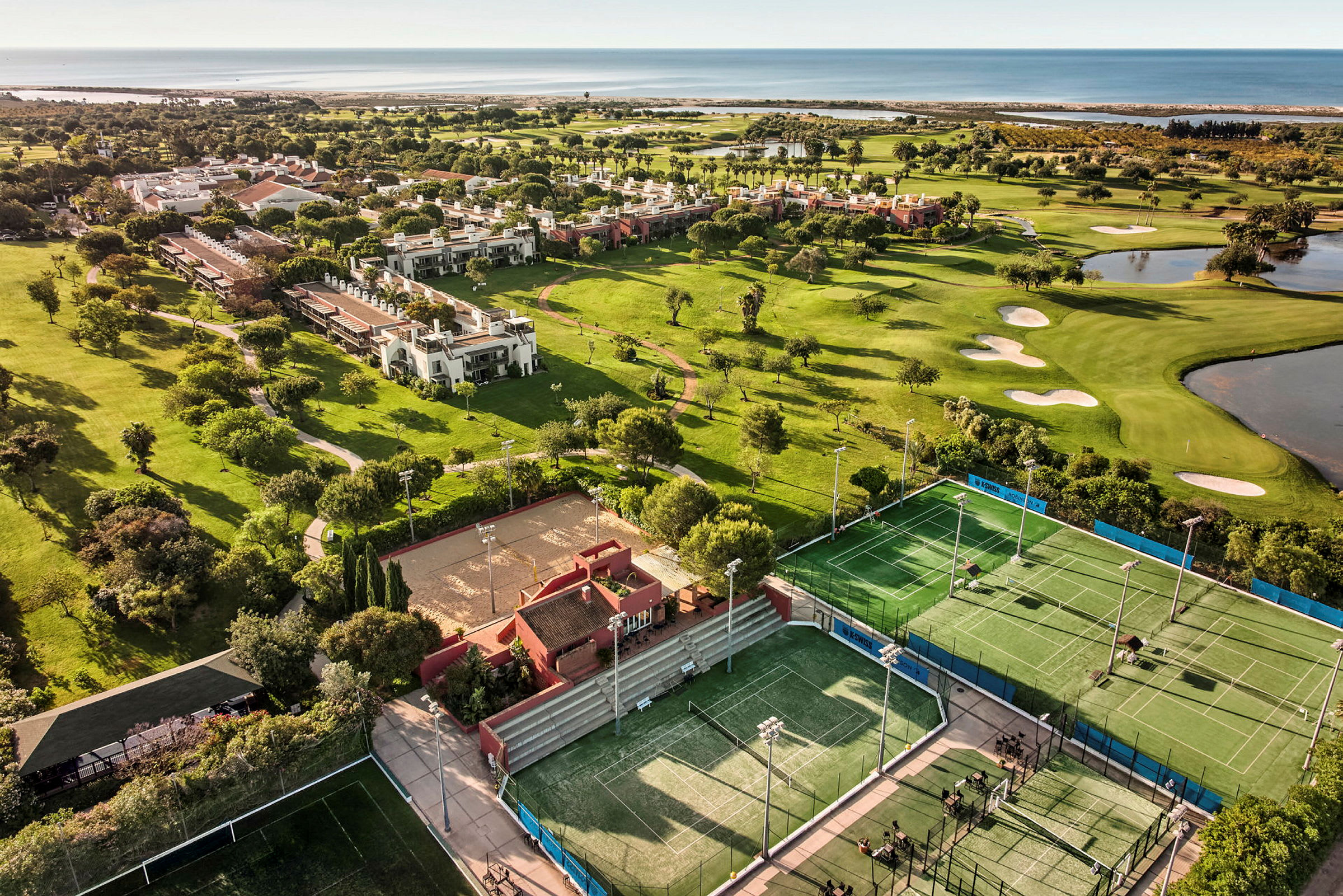 Luftfoto over en resort med tennisbaner, en golfbane og nærliggende bygninger omgitt av grøntområder og nær havet. (AI-generert tekst)