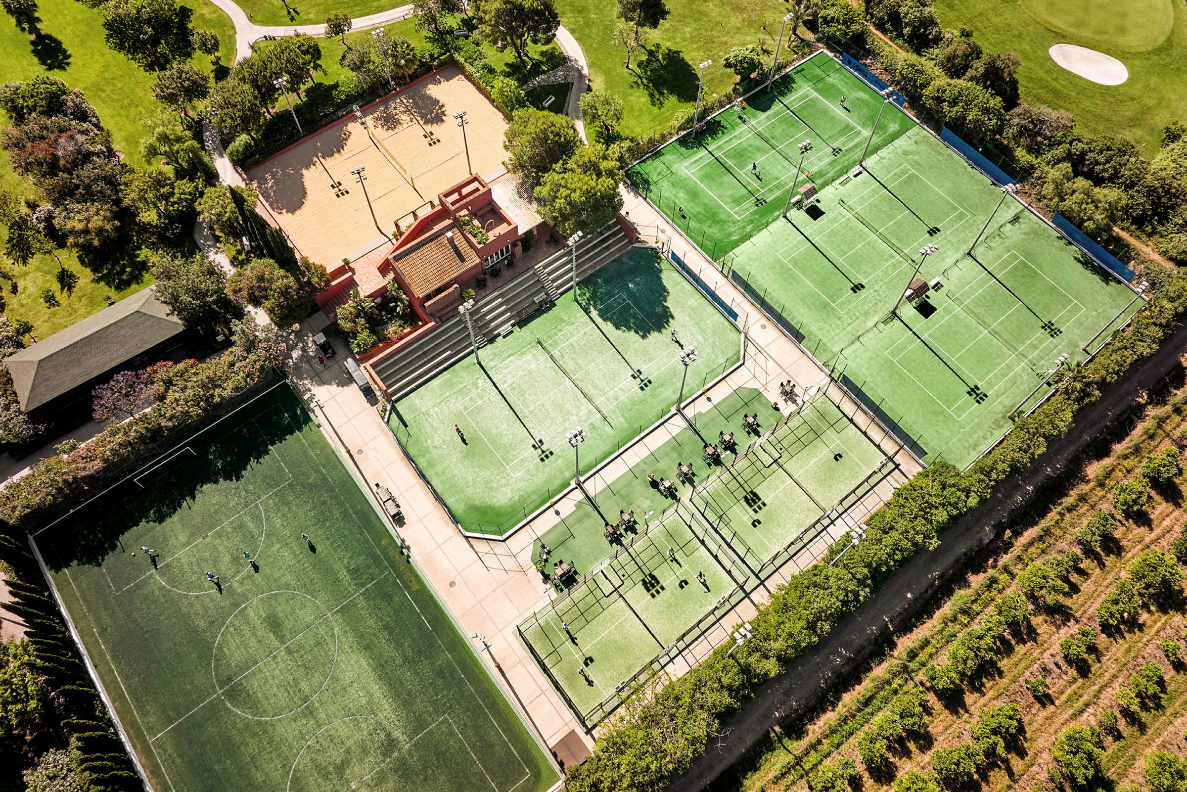 Luftfoto av et sportsanlegg med tennisbaner, padelbaner, en fotballbane og et sandfelt, omgitt av grøntområder. (AI-generert tekst)