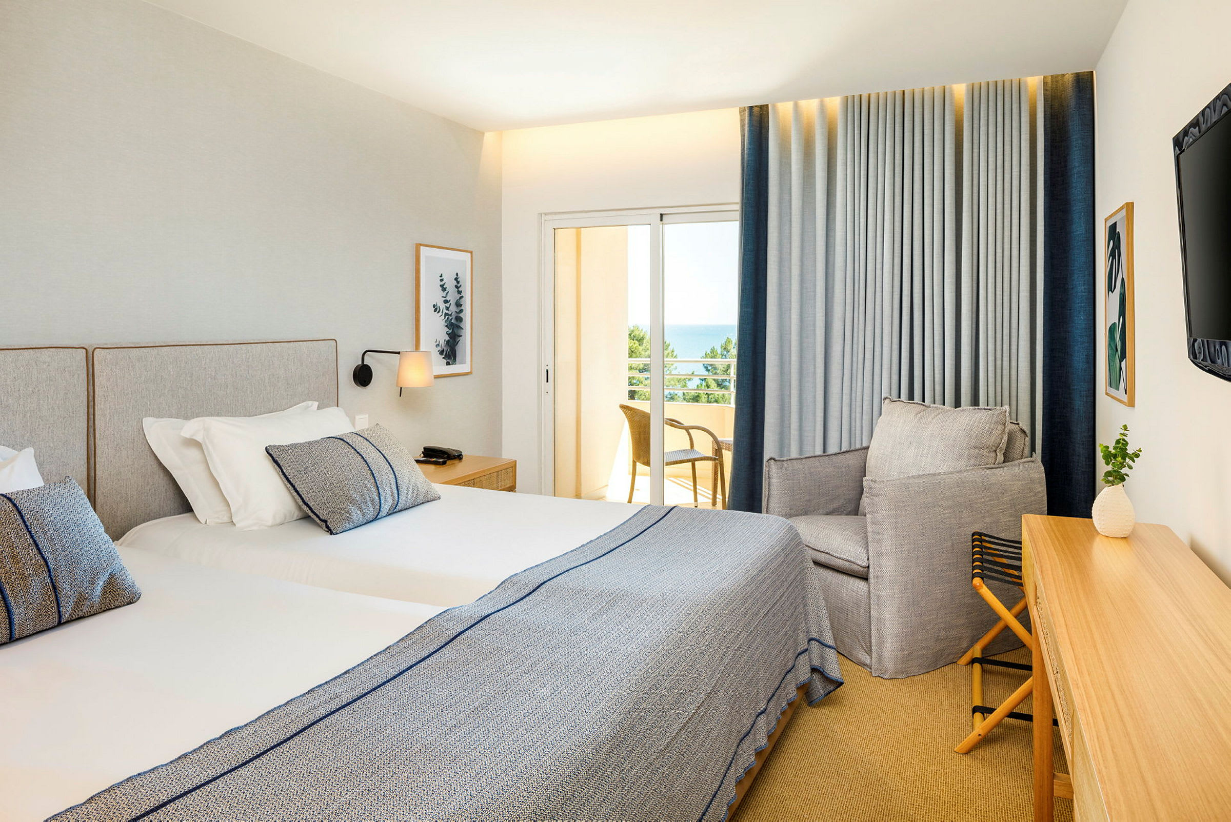 Et hotellrom med to enkeltsenger, en lenestol og et skrivebord. Balkongen har utsikt over havet. (AI-generert tekst)