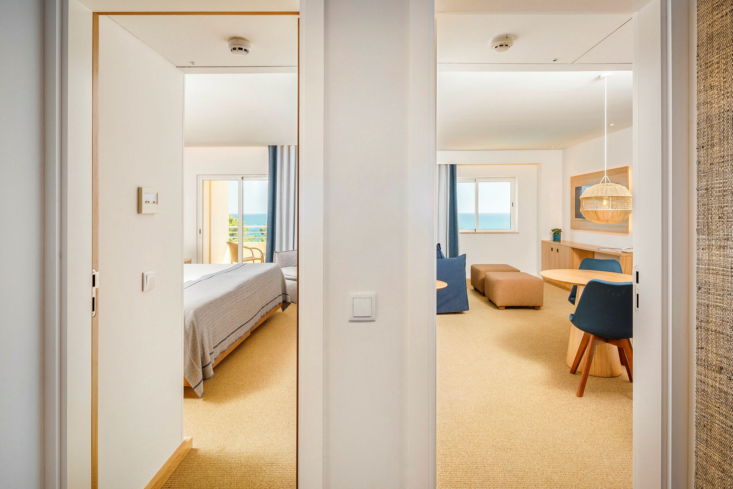Utsikt gjennom to døråpninger mot et lyst hotellrom med soveområde og sittegruppe, samt et stort vindu med havutsikt. (AI-generert tekst)