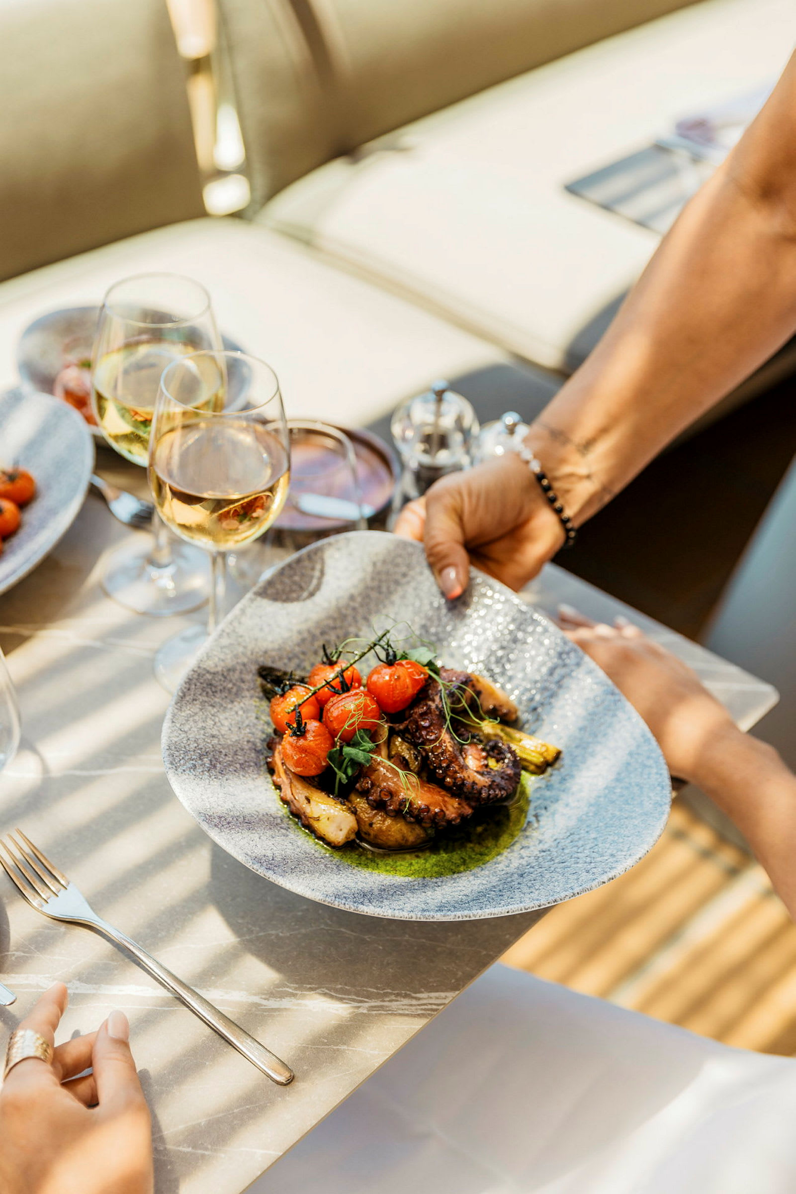 Bildet viser en tallerken med en delikat matrett bestående av grillet blekksprut, tomater og grønnsaker, servert på et bord med vinglass. (AI-generert tekst)