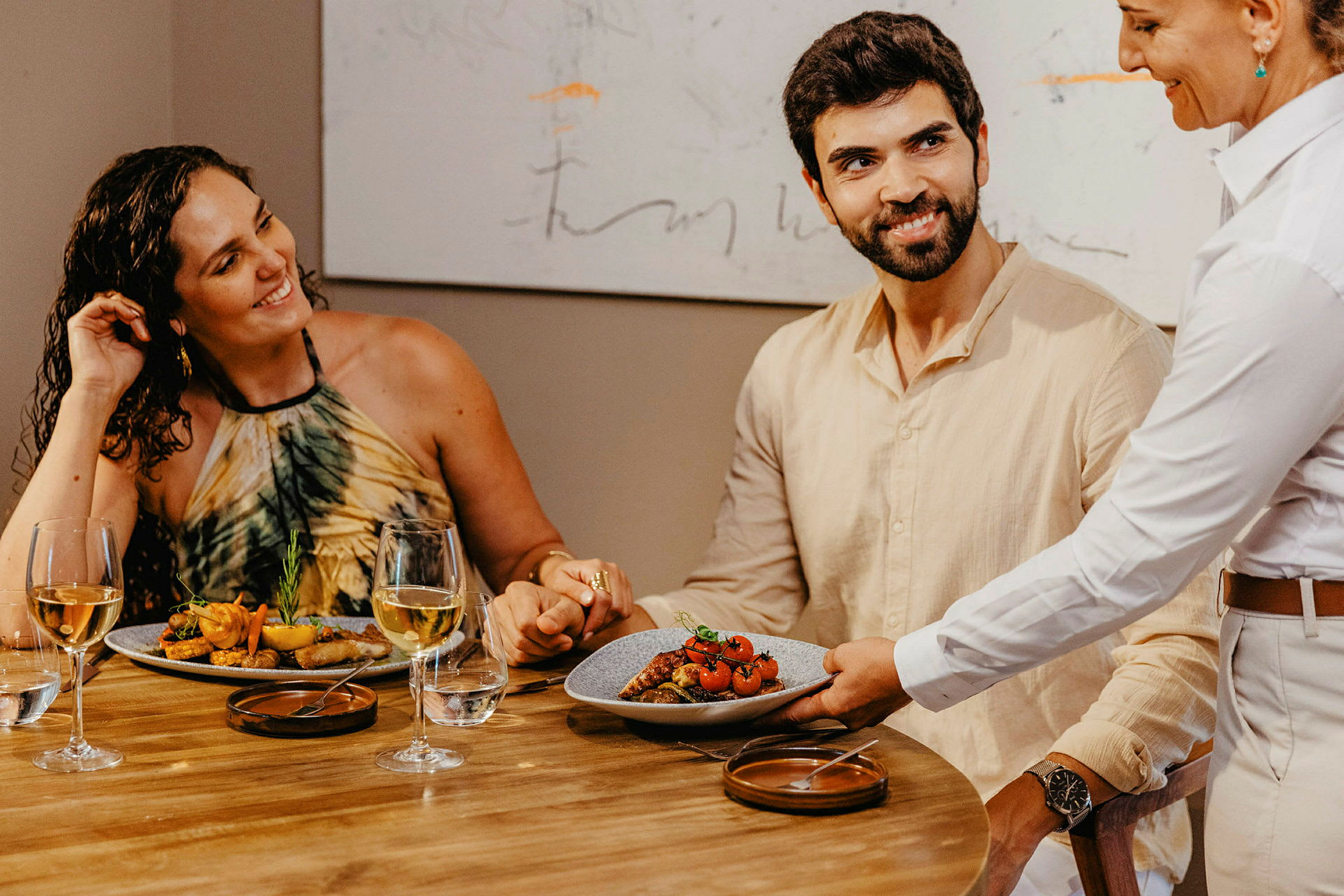 En servitør serverer en tallerken med mat på et bord hvor to personer sitter og holder hender. (AI-generert tekst)