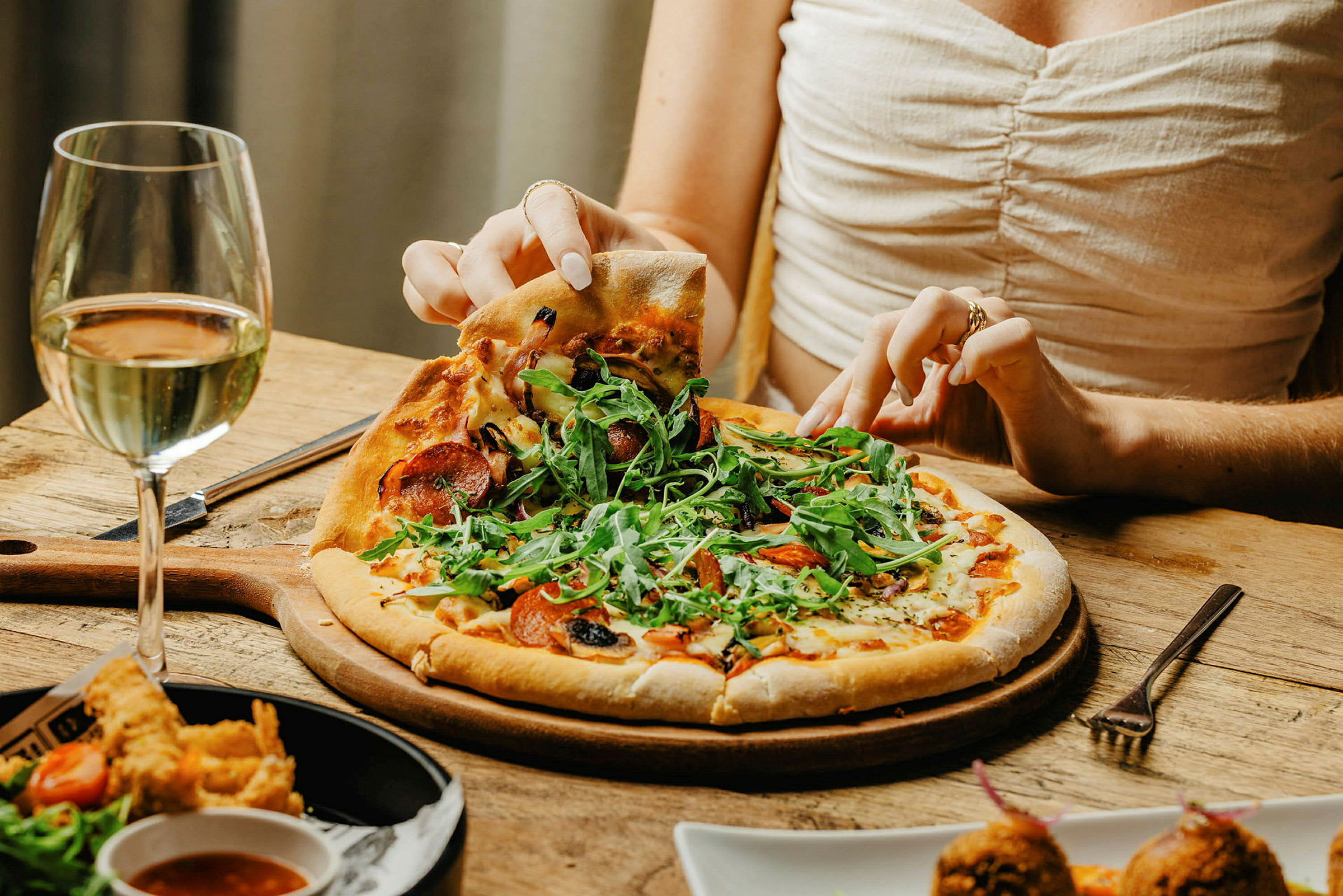 Nærbilde av en person som tar et stykke pizza fra en tallerken med et glass hvitvin ved siden av. (AI-generert tekst)