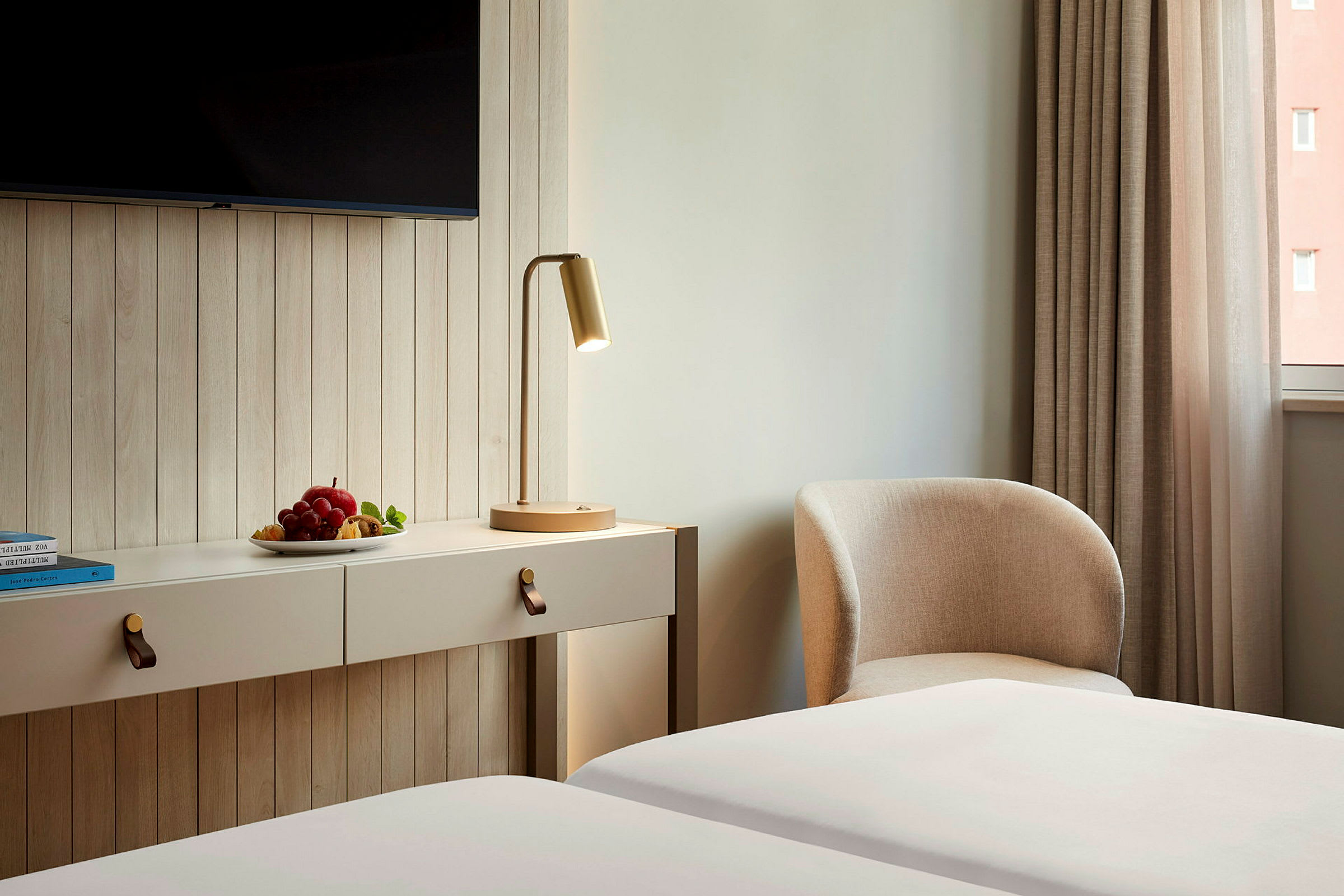 Et hotellrom med en veggmontert møbel, en bordlampe og en lenestol ved siden av en gardin samt en tallerken med frukt. (AI-generert tekst)