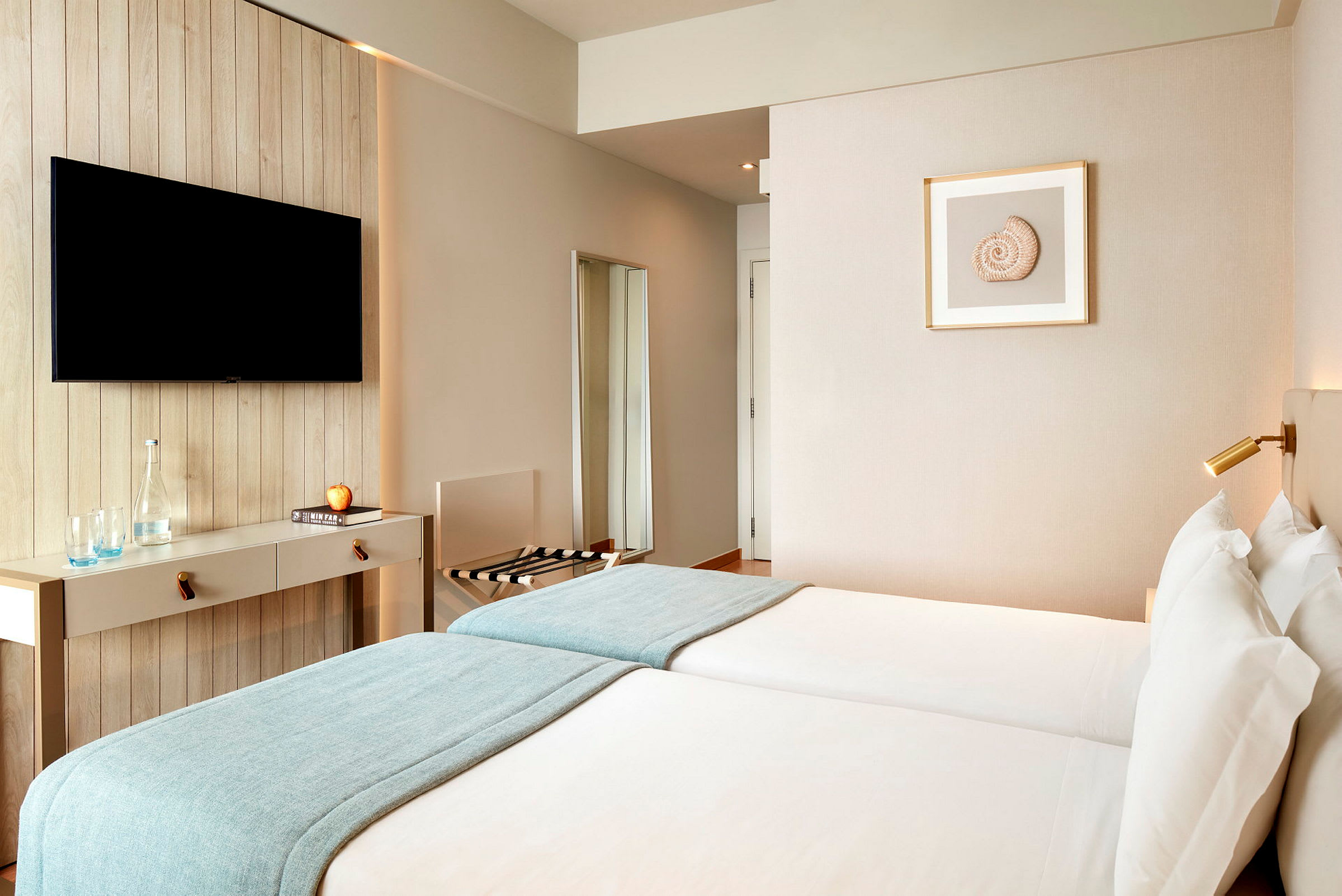 Et hotellrom med to enkeltsenger, en veggmontert TV og minimalistisk innredning. (AI-generert tekst)