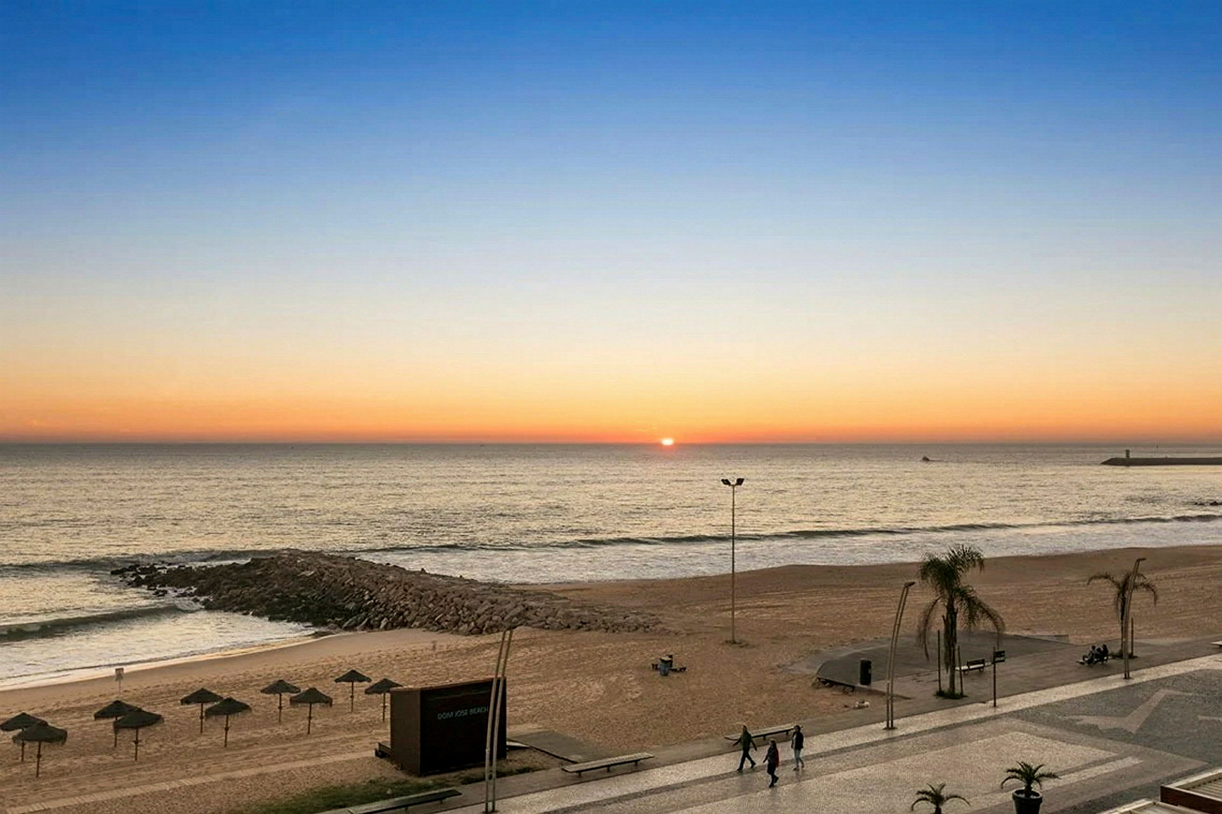 Solnedgang over en strand med parasoller og gangvei. (AI-generert tekst)