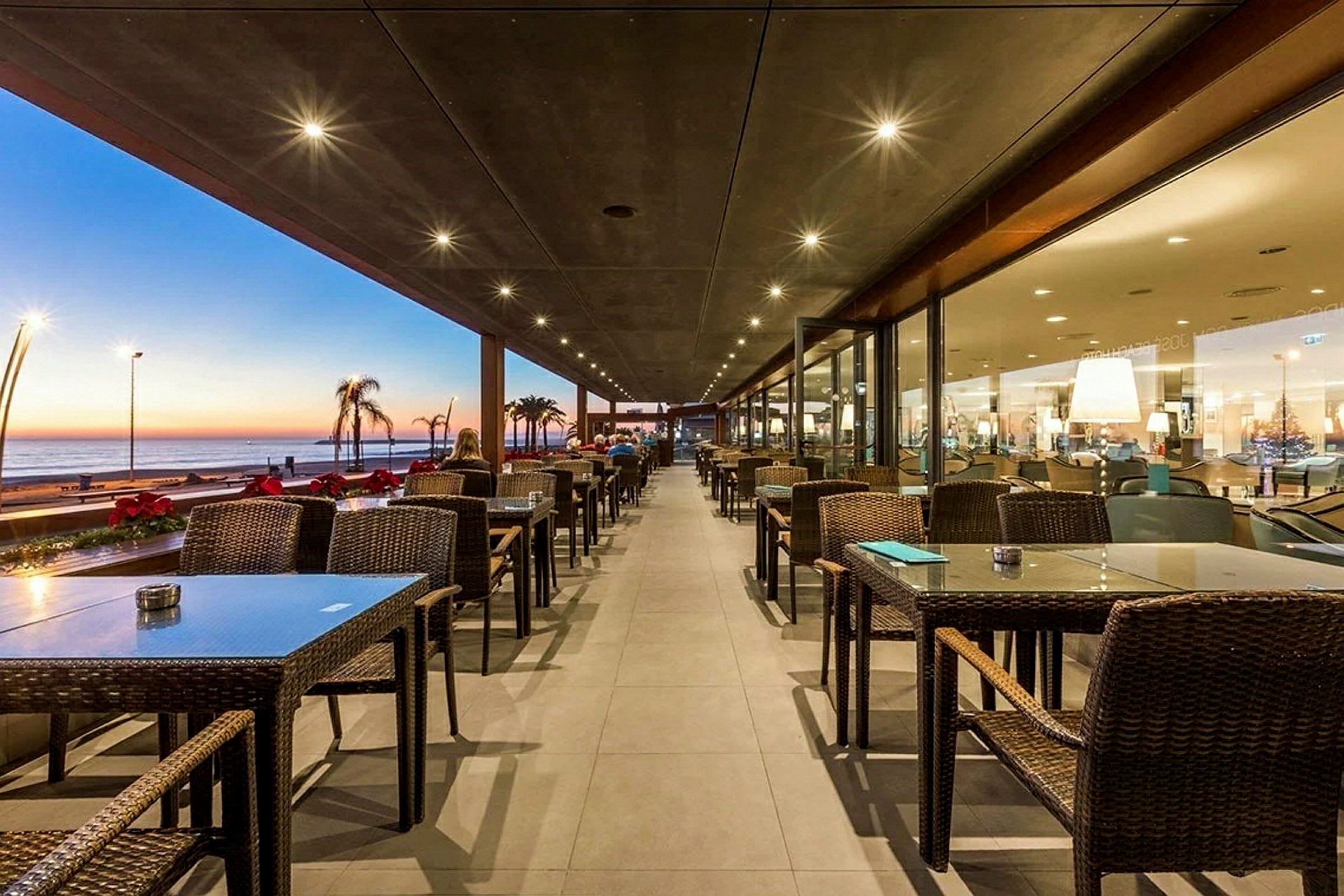Utendørsrestaurant med oppstilte bord og stoler, samt utsikt over havet under solnedgang. (AI-generert tekst)