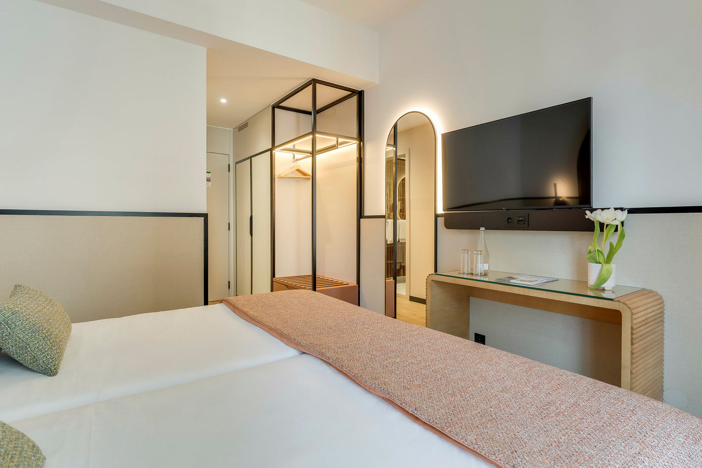 Et hotellrom med en dobbeltseng, et speil, en TV og et skrivebord med en vase med hvite blomster. (AI-generert tekst)