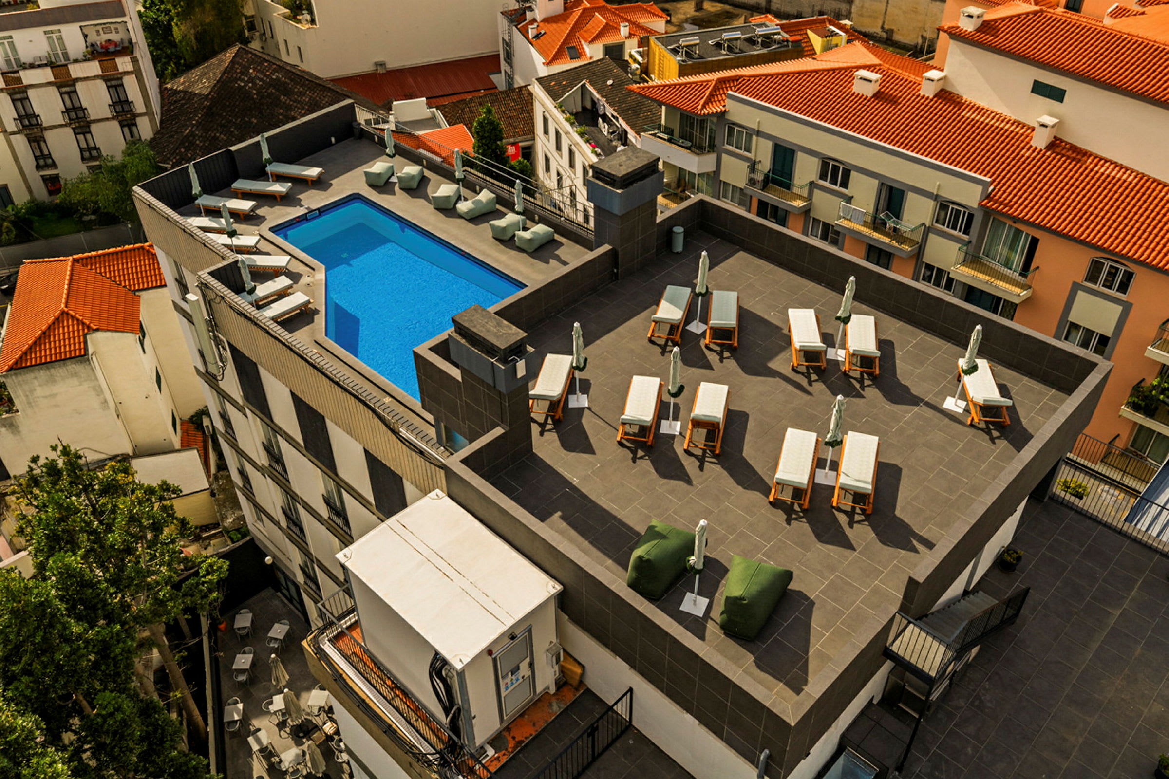 Bilde av en takterrasse med et blått basseng, solsenger og parasoller, omgitt av bygninger med tegltak. (AI-generert tekst)