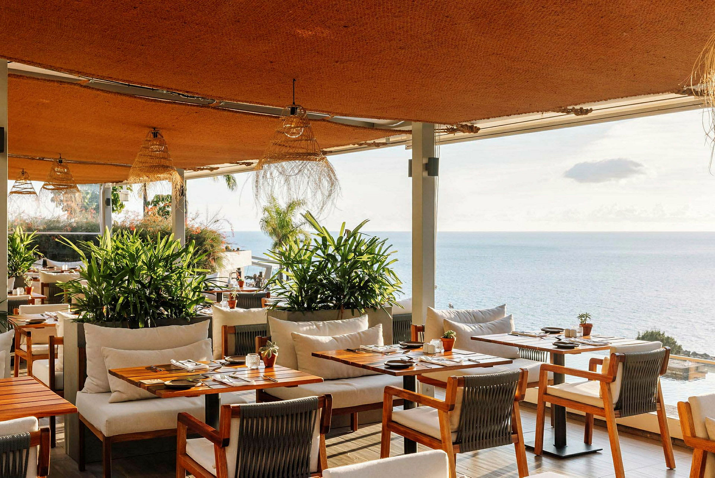 Utsikt over en restaurant med trebord og stoler, grønne planter og havet i bakgrunnen. (AI-generert tekst)