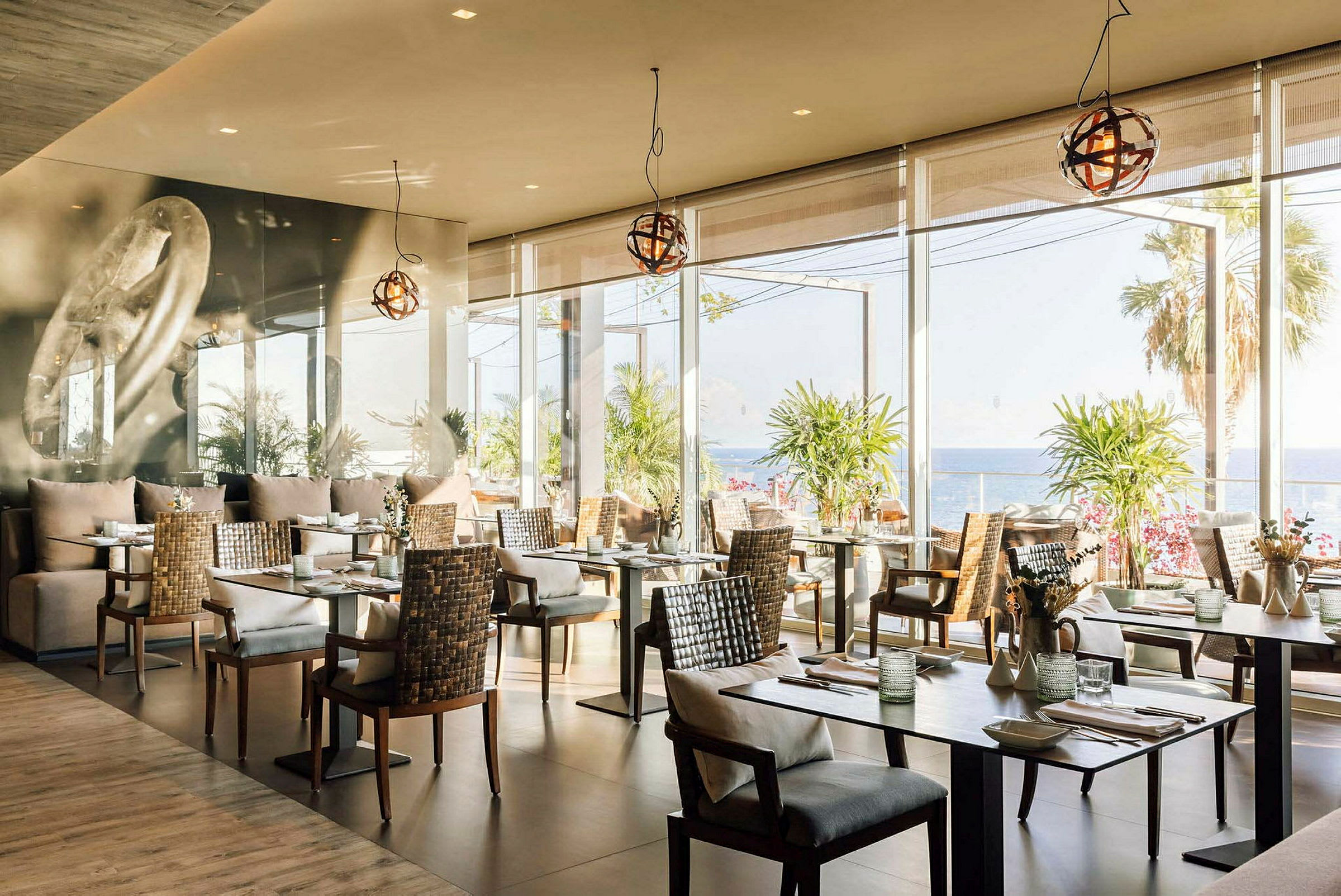 En restaurant med moderne bord og stoler, store vinduer som viser utsikt over havet og palmetrær. (AI-generert tekst)