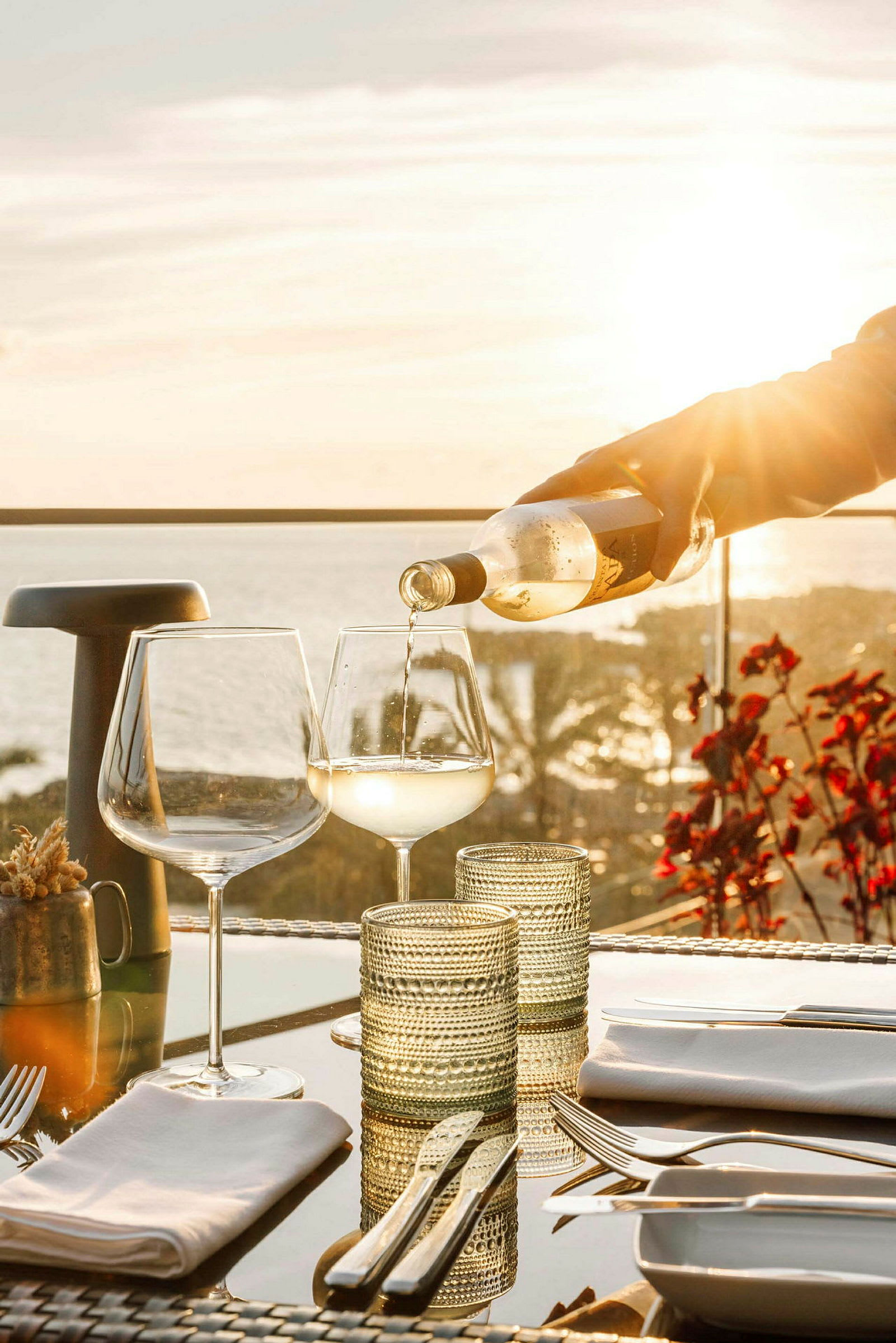 Hvit drikke helles i et vinglass på et bord med redskaper og glass, med solnedgang og hav i bakgrunnen. (AI-generert tekst)