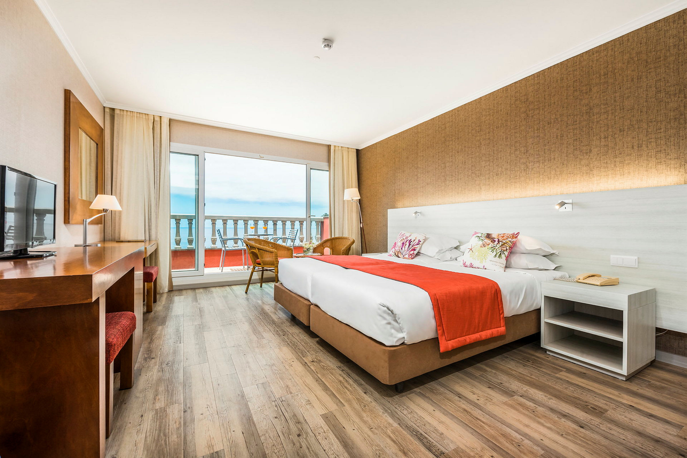 Hotellrom med to senger, et bord og utsikt over en balkong og havet. (AI-generert tekst)
