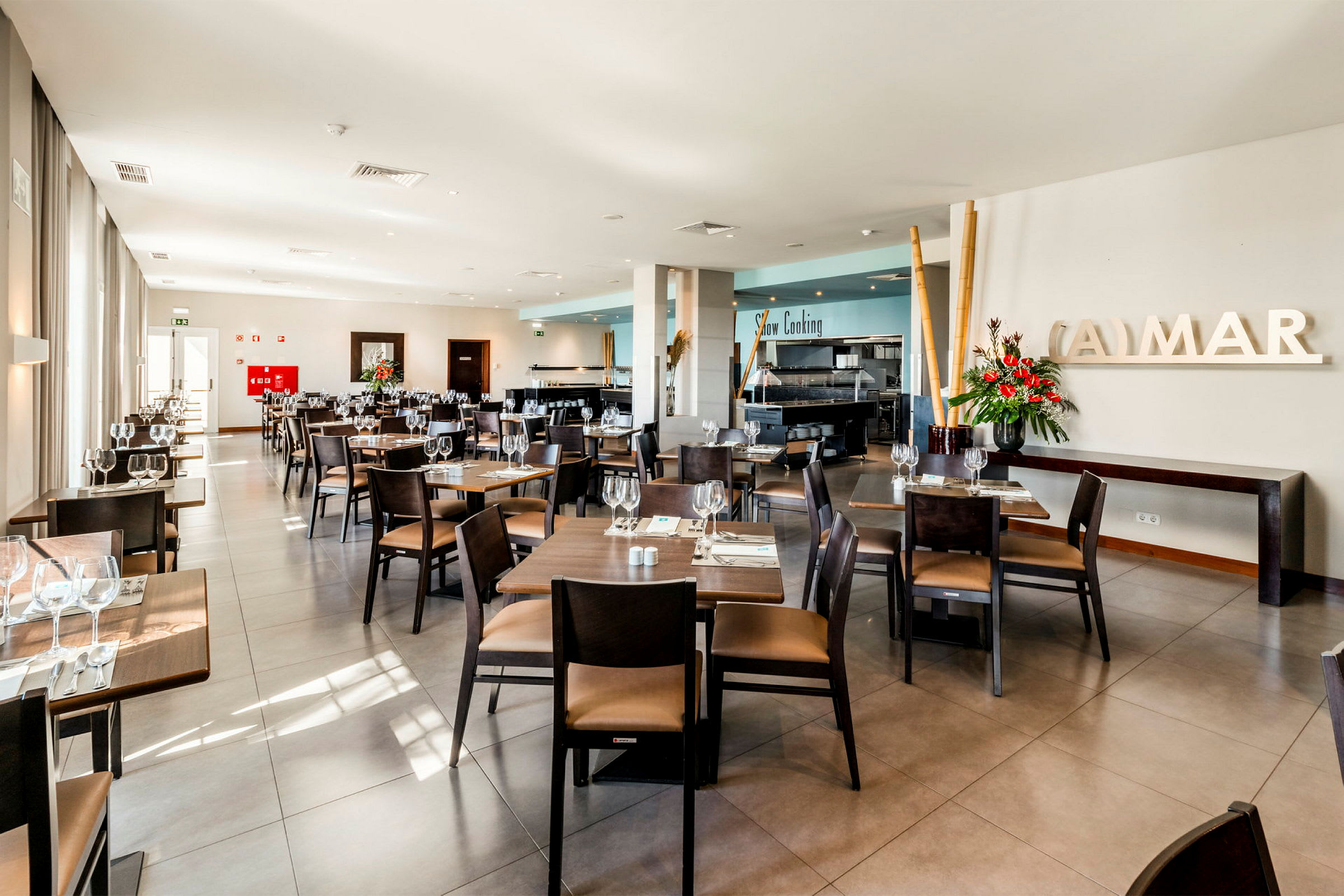 En lys og moderne restaurant med bord og stoler pent arrangert, friske blomster og store vinduer. (AI-generert tekst)