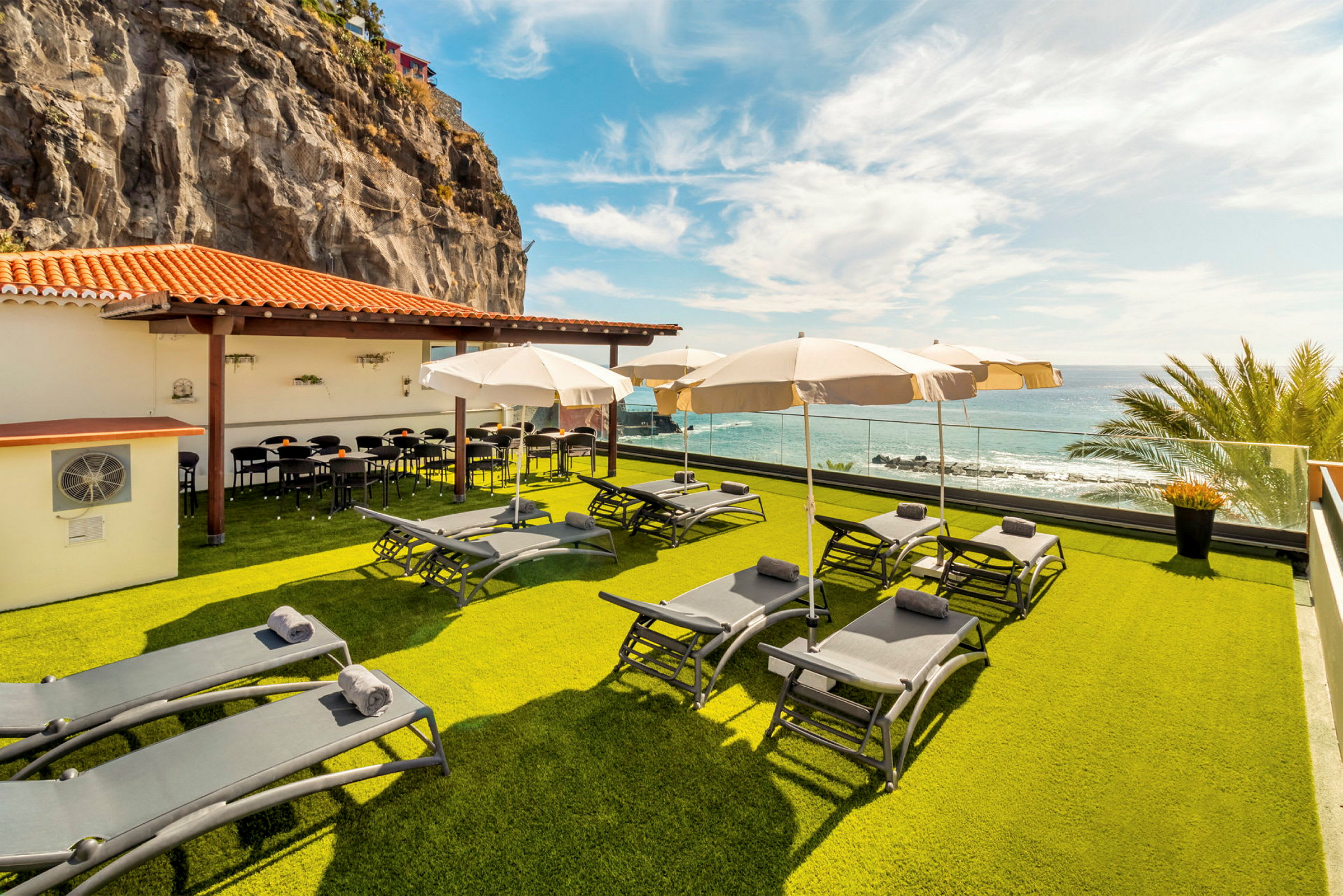 En terrasse med solsenger og parasoller på en gressplen, ved et lavt hus med tak i terrakottarødt. Utsikt over havet og en klippevegg i bakgrunnen. (AI-generert tekst)