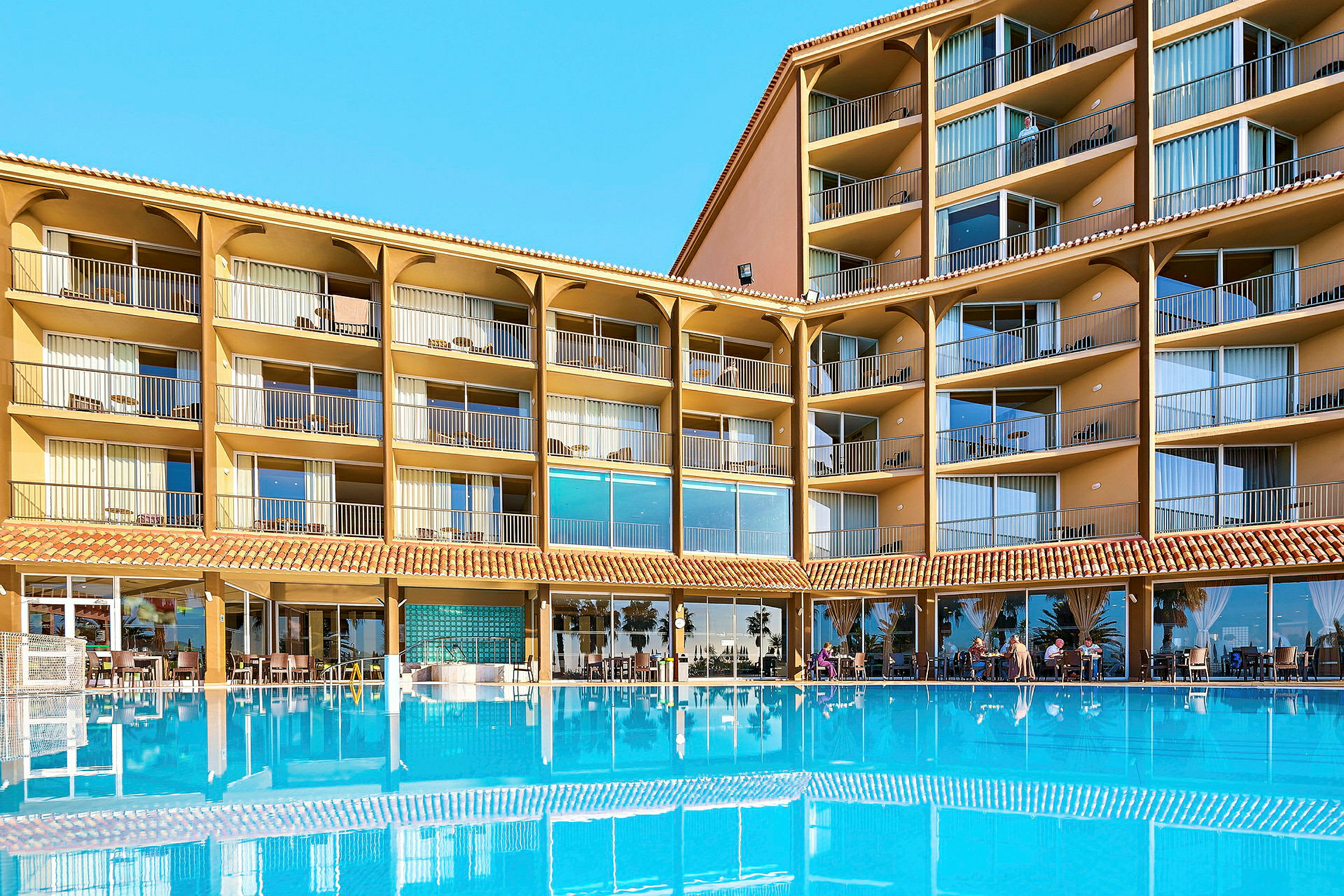 Hotellbygning med flere etasjer og balkonger, foran et stort blått basseng. (AI-generert tekst)