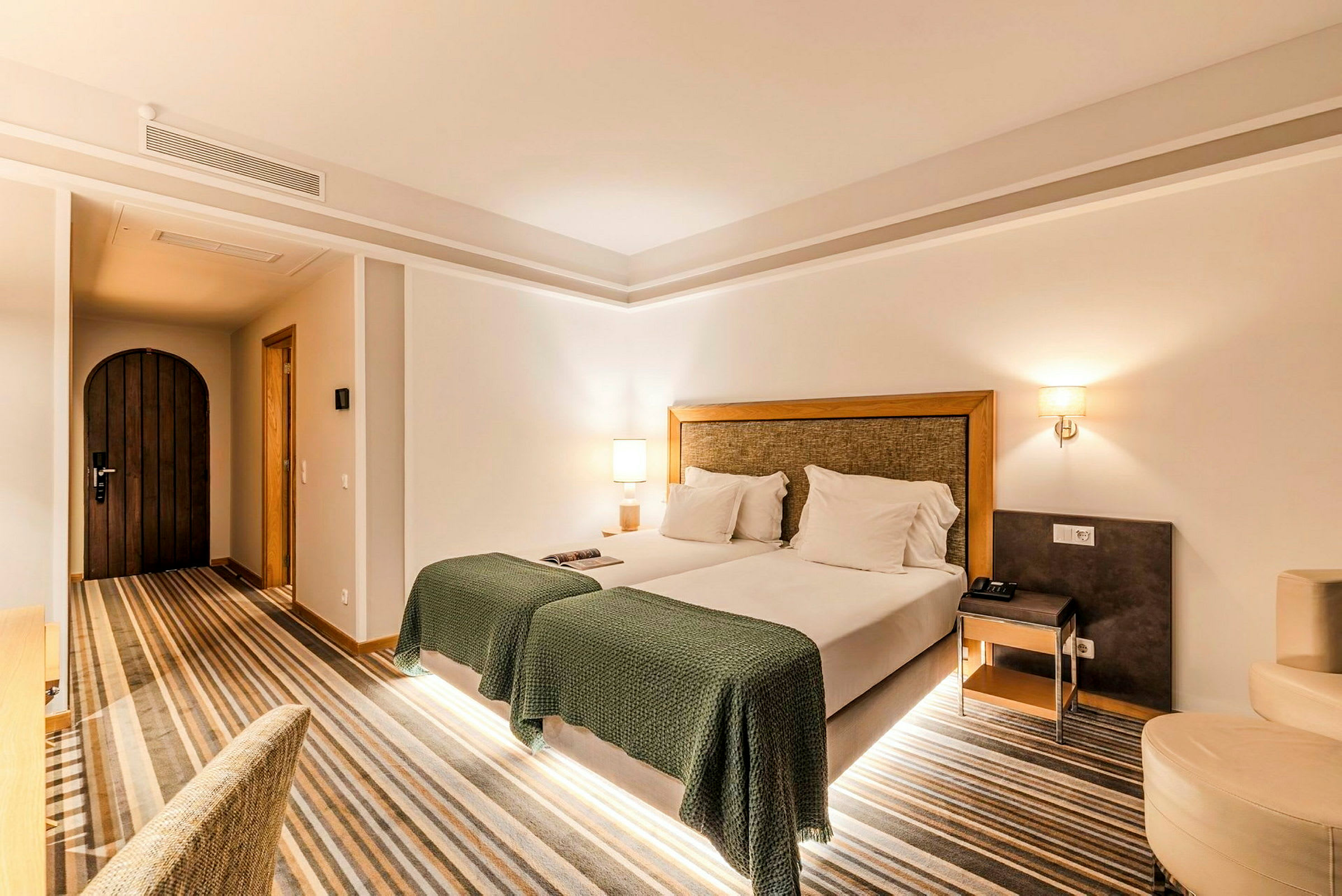 Et hotellrom med en dobbeltseng, grønne sengetepper og to hvite lamper på nattbordene på hver side. En tredør er synlig på den ene veggen. (AI-generert tekst)