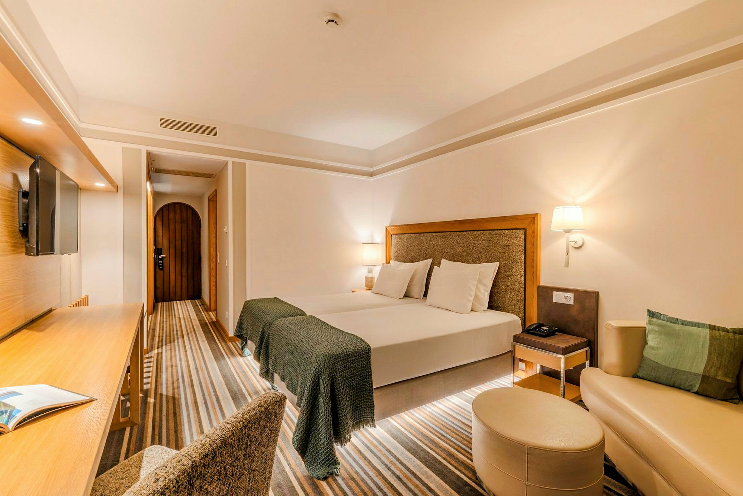 Et godt opplyst hotellrom med en stor dobbeltseng, skrivebord, stol, veggmontert TV og en liten sittegruppe med sofa og fotskammel. (AI-generert tekst)