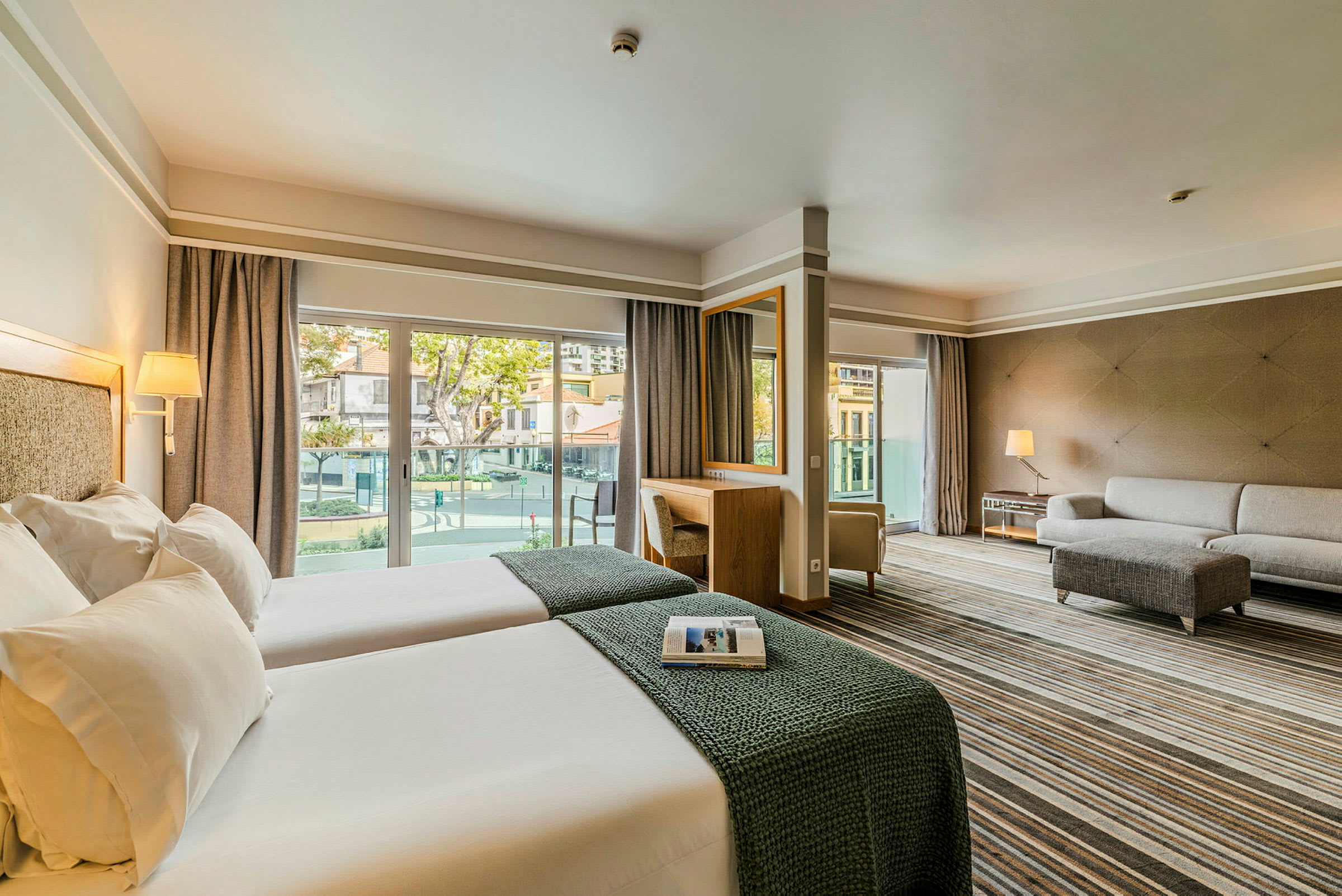 Et hotellrom med to enkeltsenger, en sofa og et skrivebord samt vinduer som fører ut til en balkong. (AI-generert tekst)