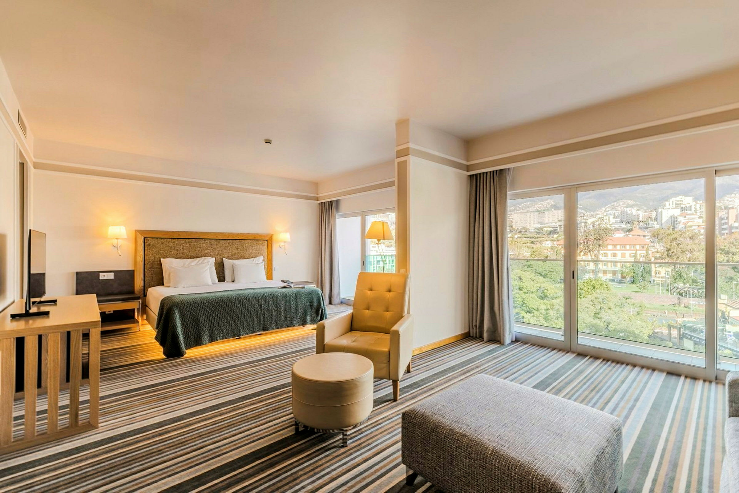 Et hotellrom med en stor seng, lenestol og sofabord samt store vinduer med utsikt over en by. (AI-generert tekst)