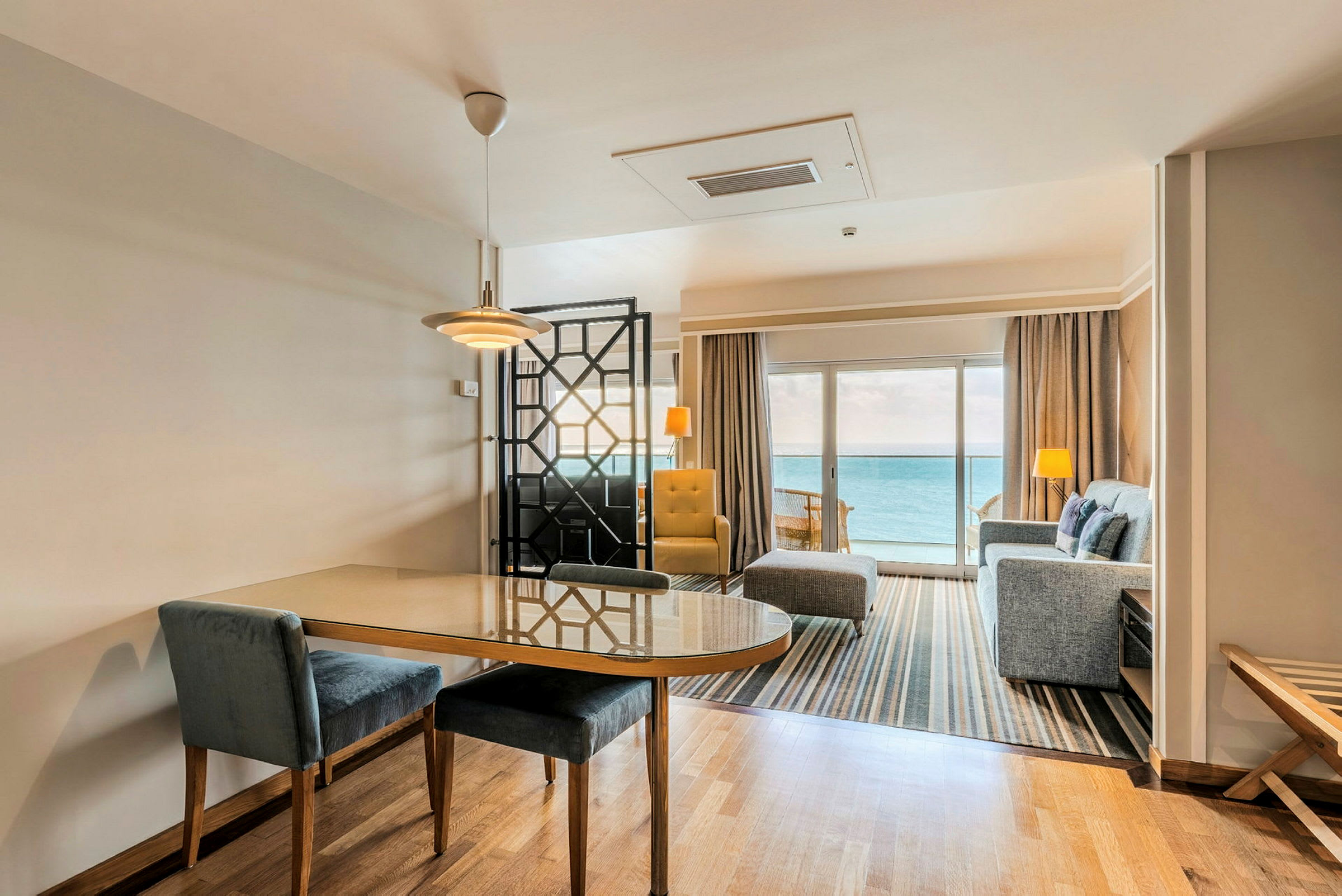 Et hotellrom med sittegruppe og spisebord, med utsikt over havet fra en stor glassdør. (AI-generert tekst)