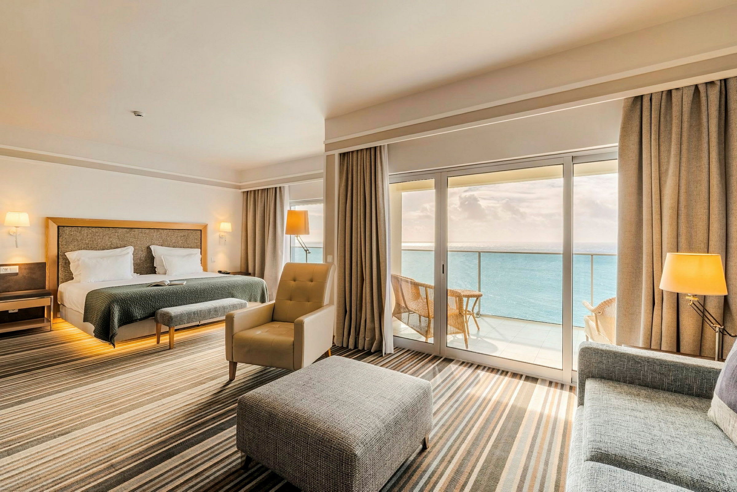 Et hotellrom med dobbeltseng, sitteområde og store vinduer som vender mot en balkong med utsikt over havet. Rommet har en moderne og stilig innredning. (AI-generert tekst)