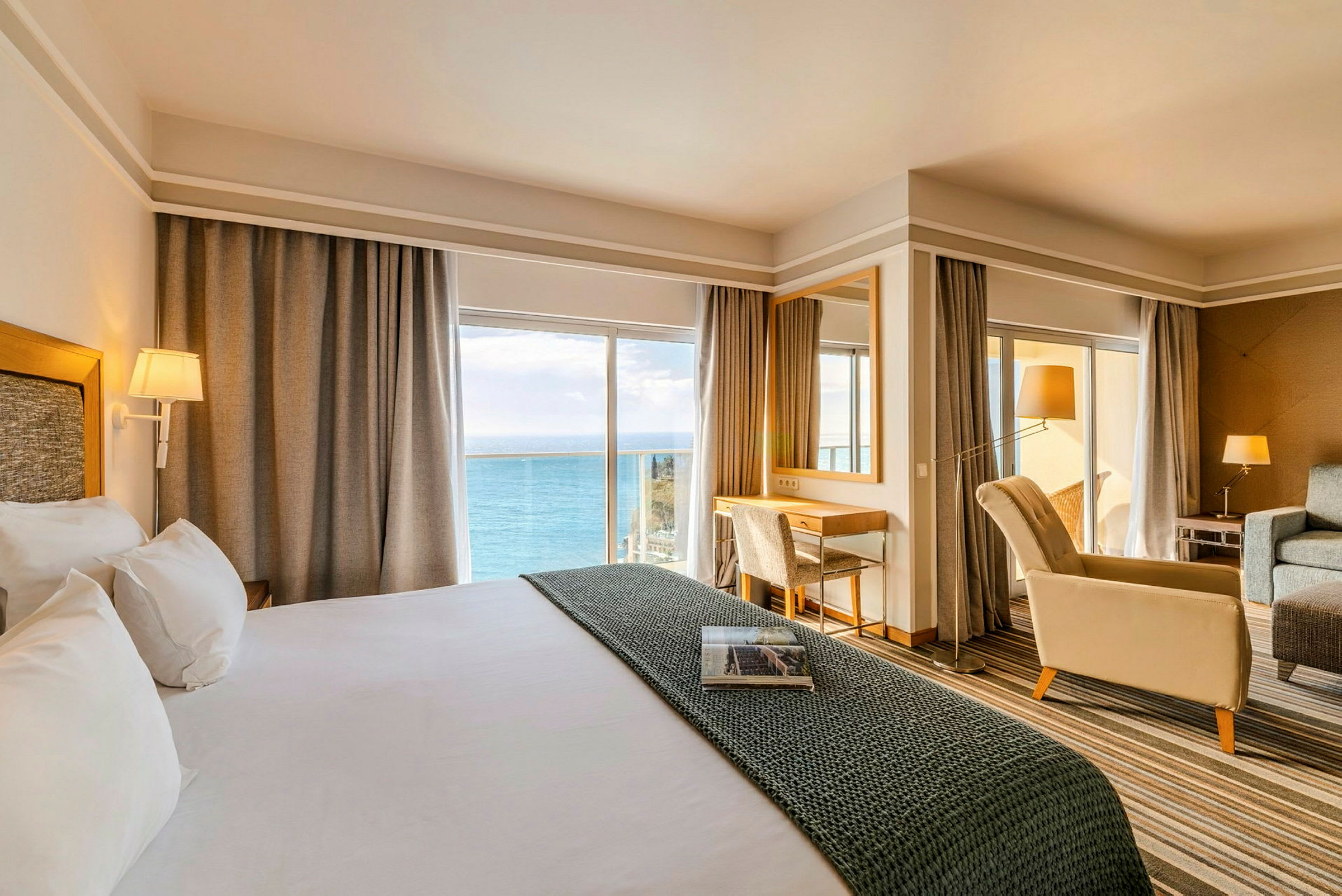Et hotellrom med stor seng, sittegruppe og skrivebord, med utsikt over havet gjennom store vinduer. (AI-generert tekst)