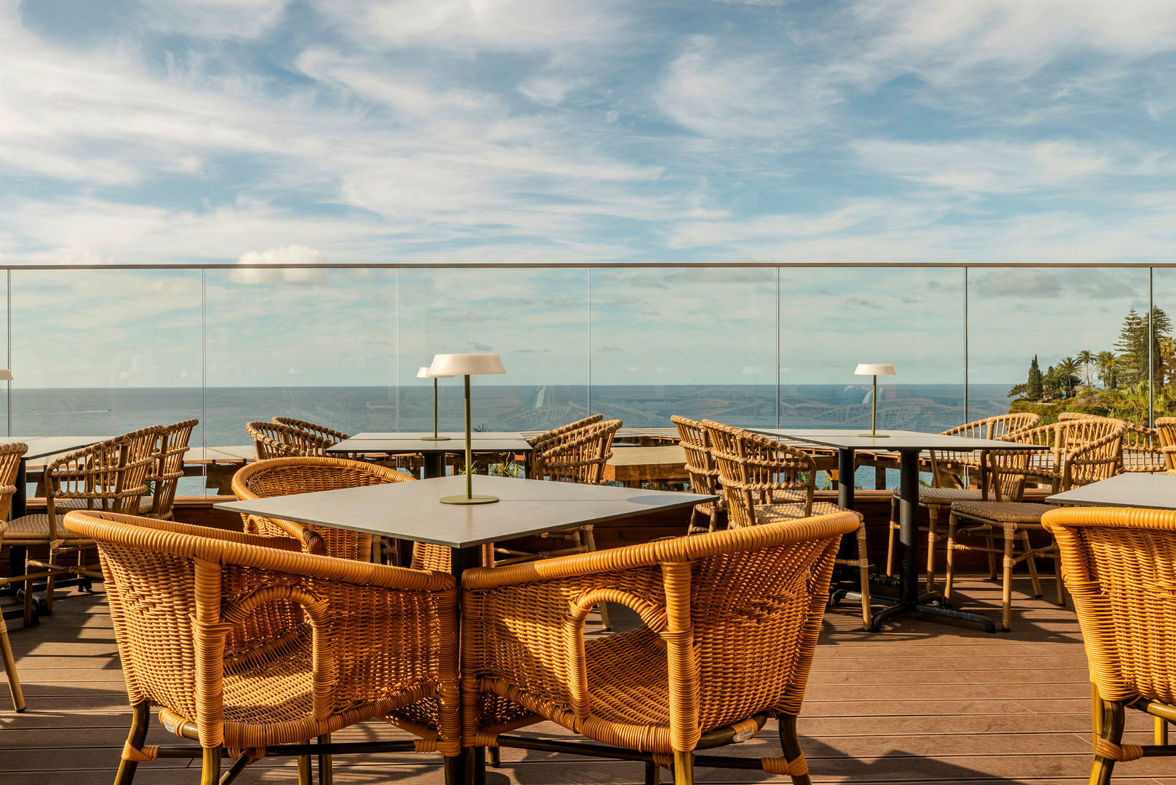 En terrasse med bord og rottingstoler med utsikt over havet og omkringliggende landskap. (AI-generert tekst)