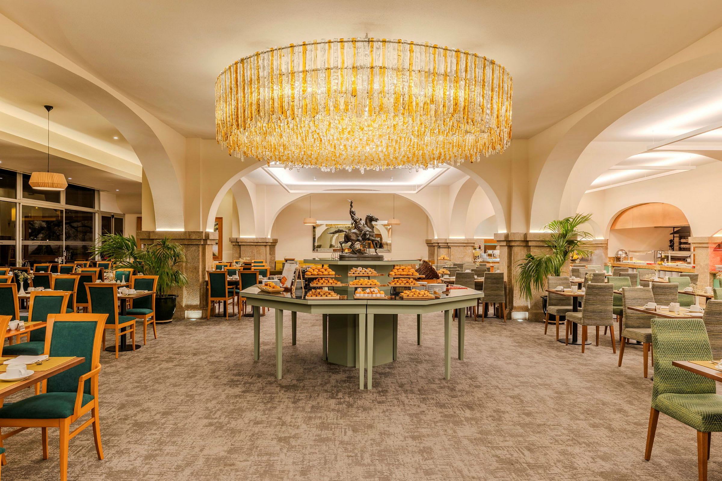 En godt opplyst hotellrestaurant med et rundt bord fullt av brød og bakverk i midten. (AI-generert tekst)
