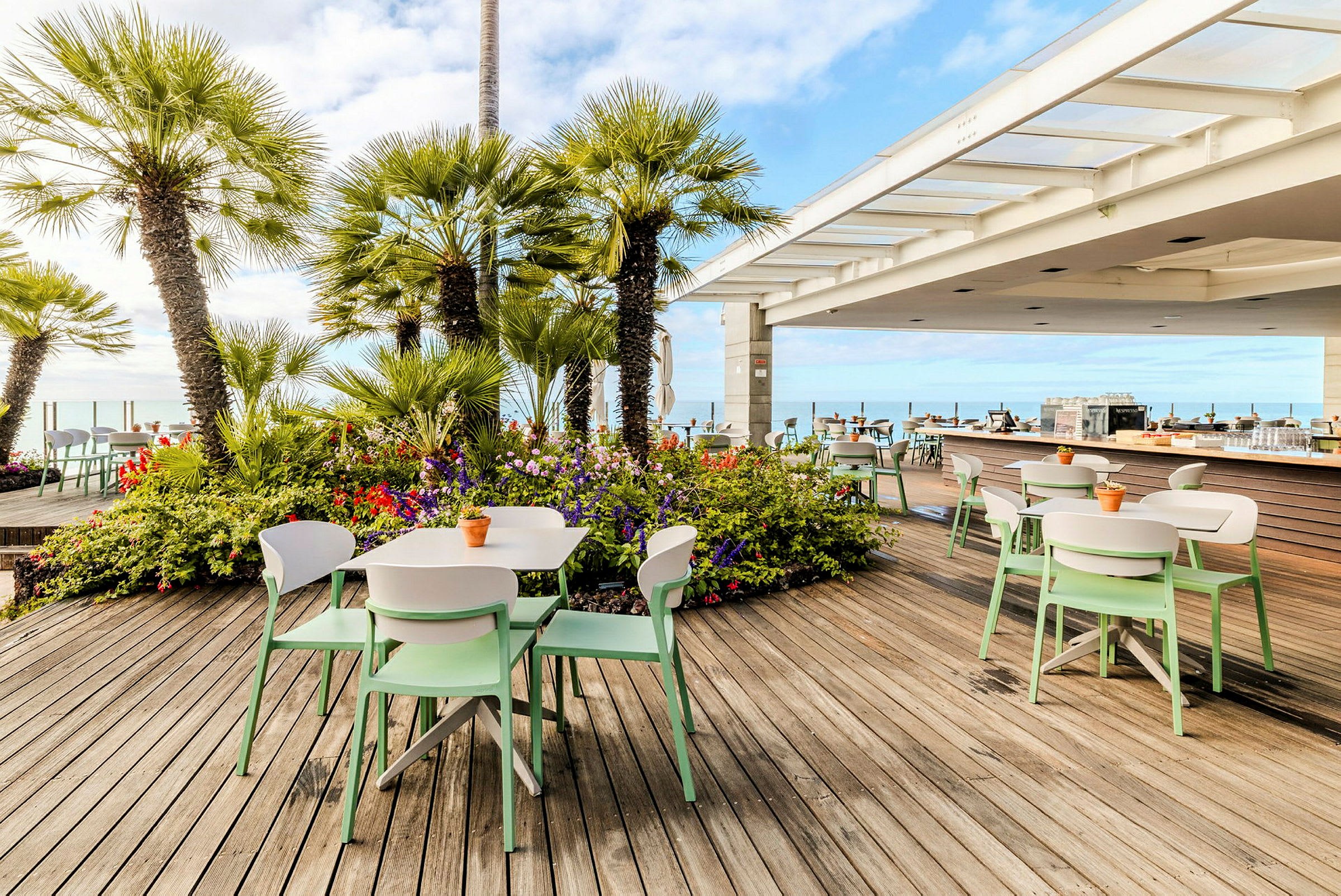 Utendørs serveringsområde på en treterrasse med grønne palmer og fargerike blomster. (AI-generert tekst)
