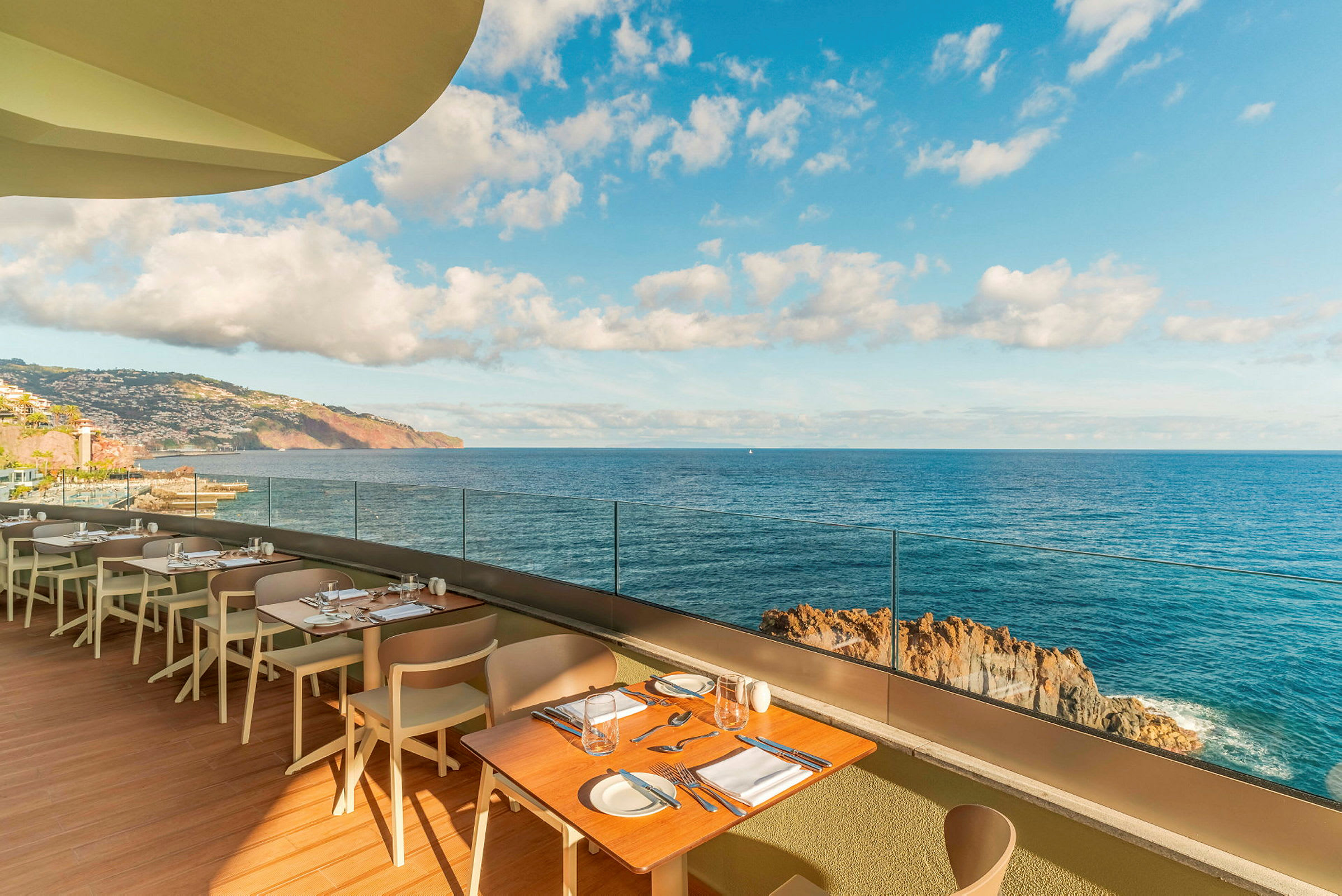 Restaurantterrasse med bord og stoler, med utsikt over havet og en klipperik kyst under en klar himmel. (AI-generert tekst)