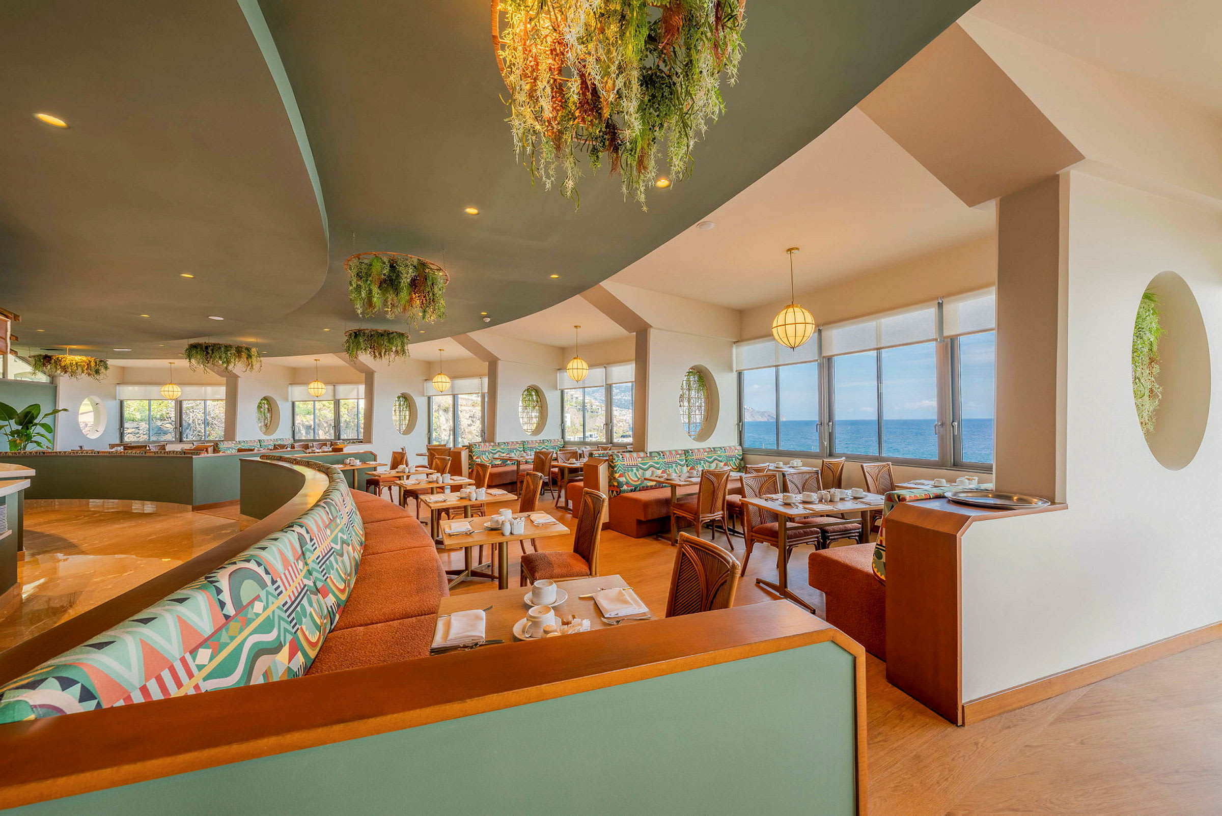 Restaurant med store vinduer og utsikt over havet, innredet med fargerike sofaer og grønne dekorasjoner. (AI-generert tekst)