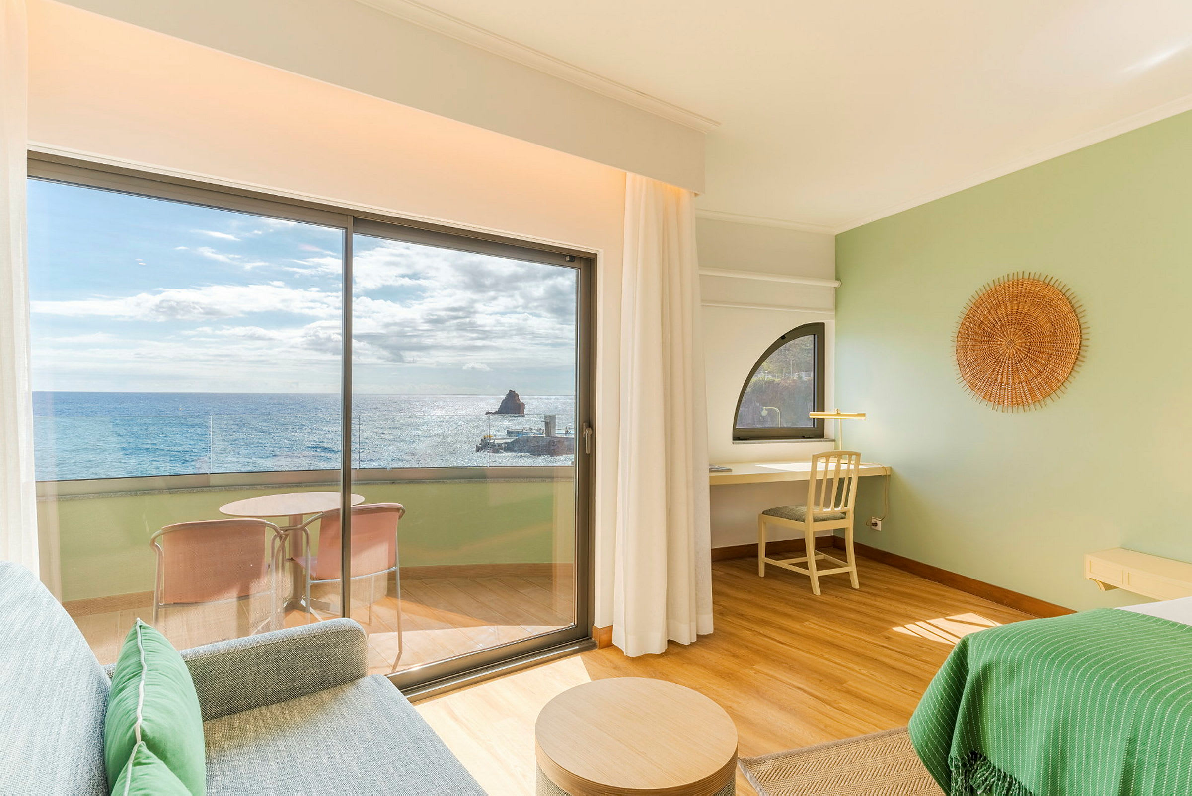 Et hotellrom med utsikt over havet gjennom store glassdører. På verandaen er det en sittegruppe, og interiøret er lyst og moderne. (AI-generert tekst)