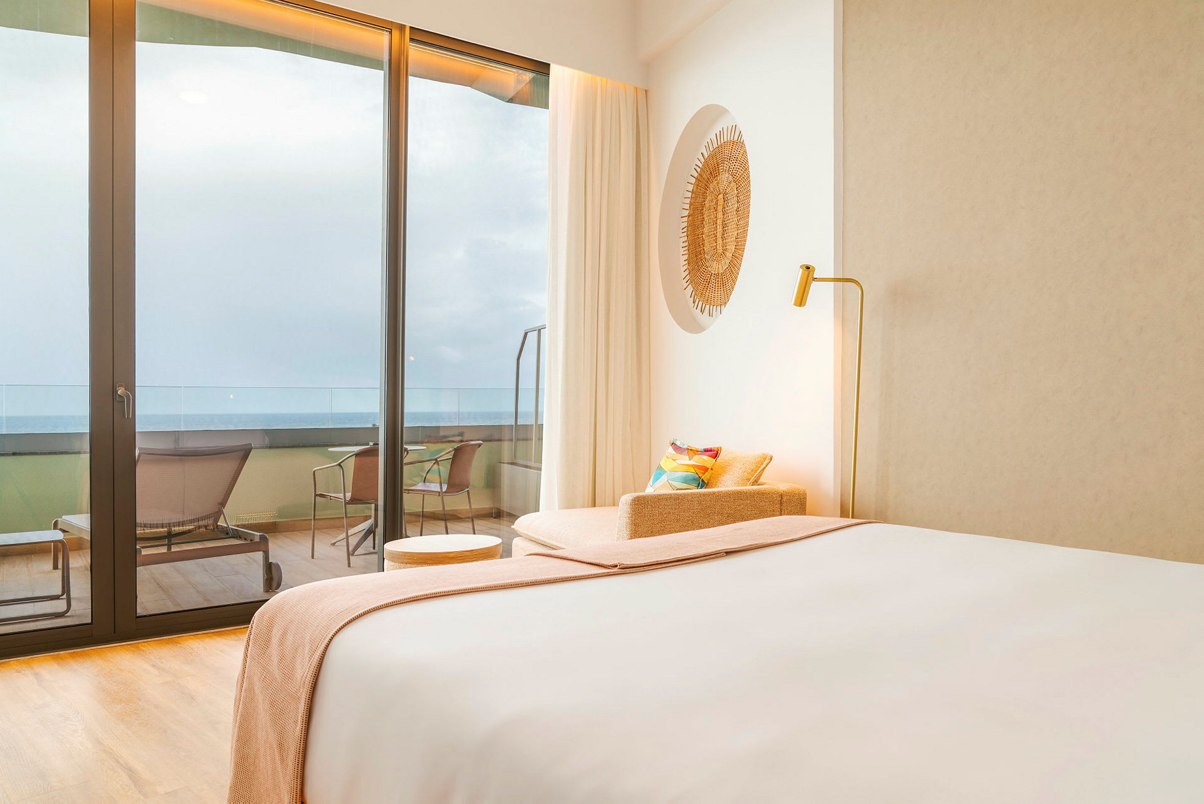 Et hotellrom med moderne interiør. Det er en seng, en lenestol og et lite bord nær en glassflate med utsikt over en balkong og havet. (AI-generert tekst)