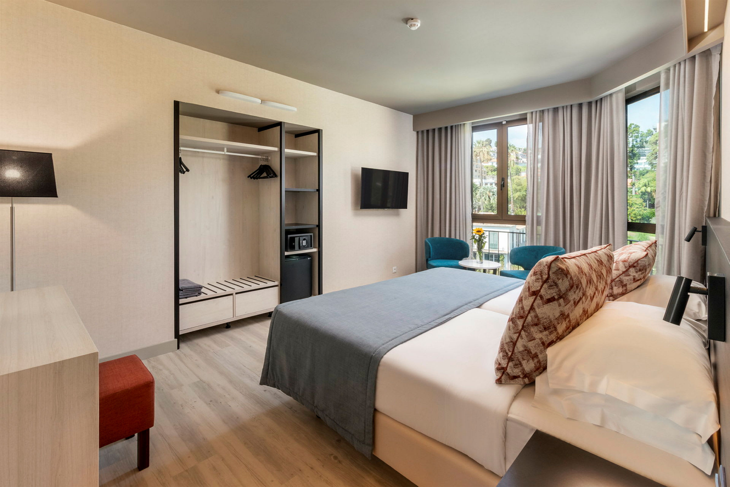 Et hotellrom med dobbeltseng, to blå stoler ved et bord foran vinduer samt garderobe med åpne hyller. (AI-generert tekst)