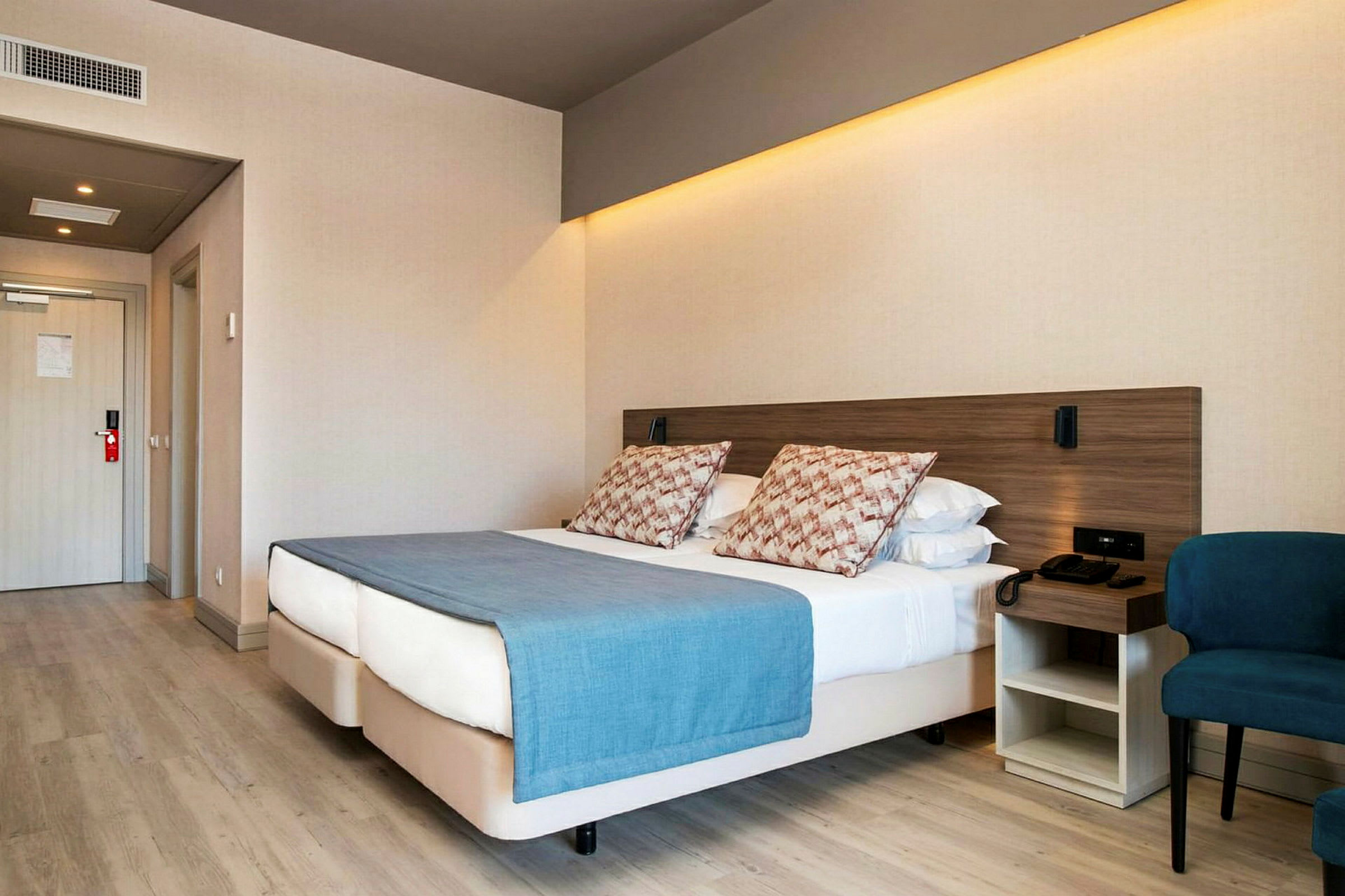 Hotellrom med en dobbeltseng, blå sengeteppe og puter, ved siden av en stol og et nattbord. (AI-generert tekst)