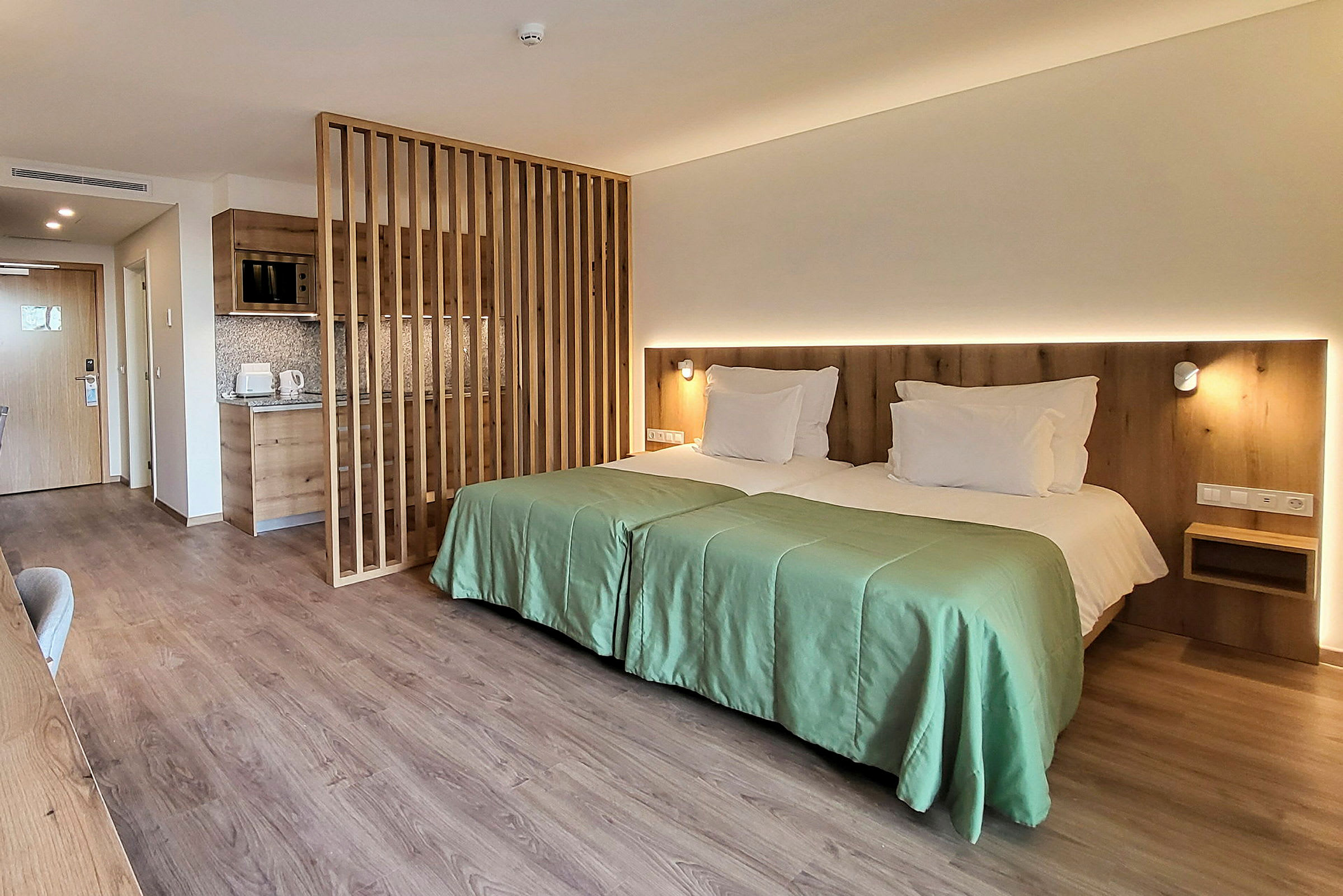 Et moderne hotellrom med to enkeltsenger ved siden av hverandre, en trepanelvegg og et lite tekjøkken. (AI-generert tekst)