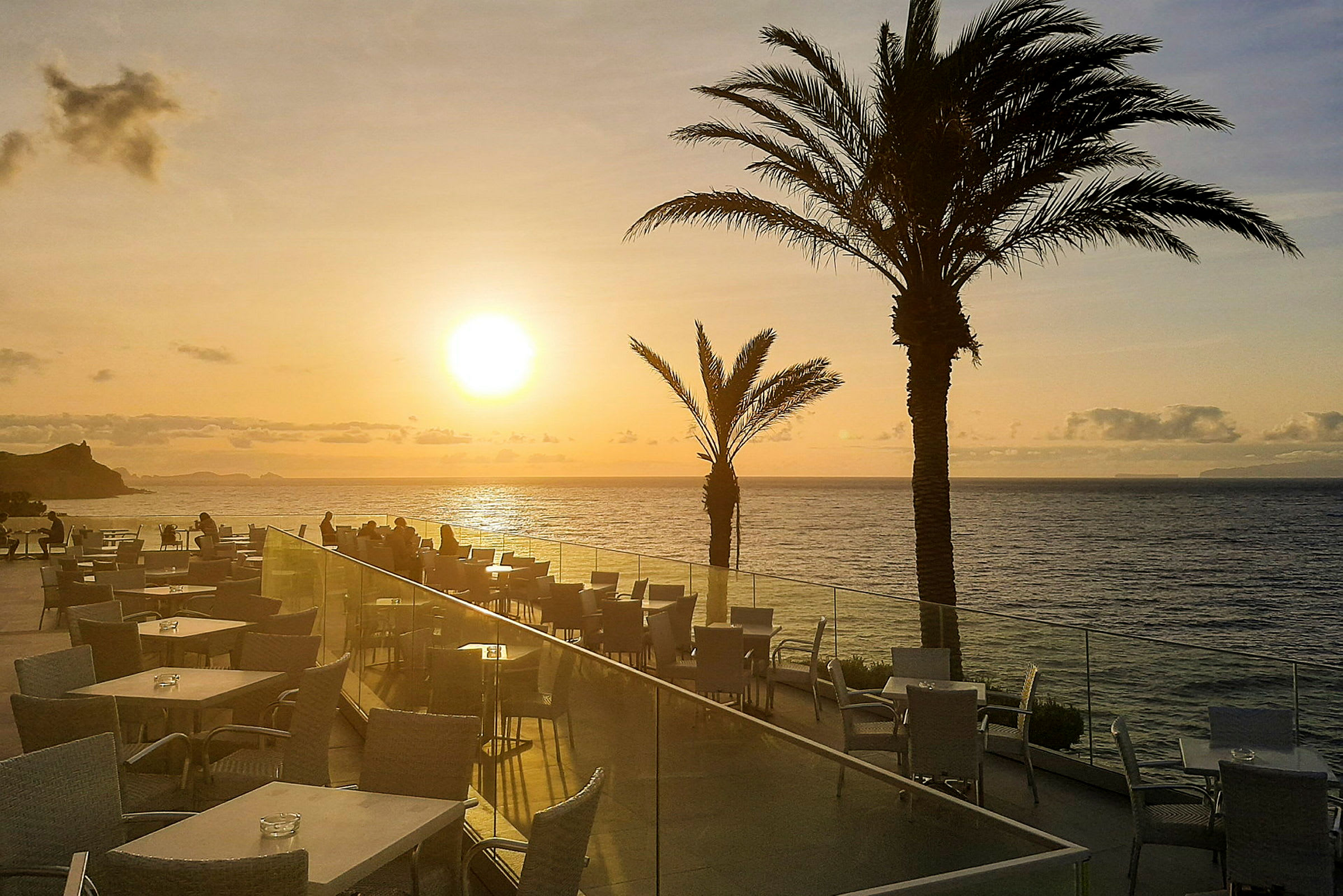 Terrasse med bord og stoler, i bakgrunnen vises havet og en solnedgang. (AI-generert tekst)
