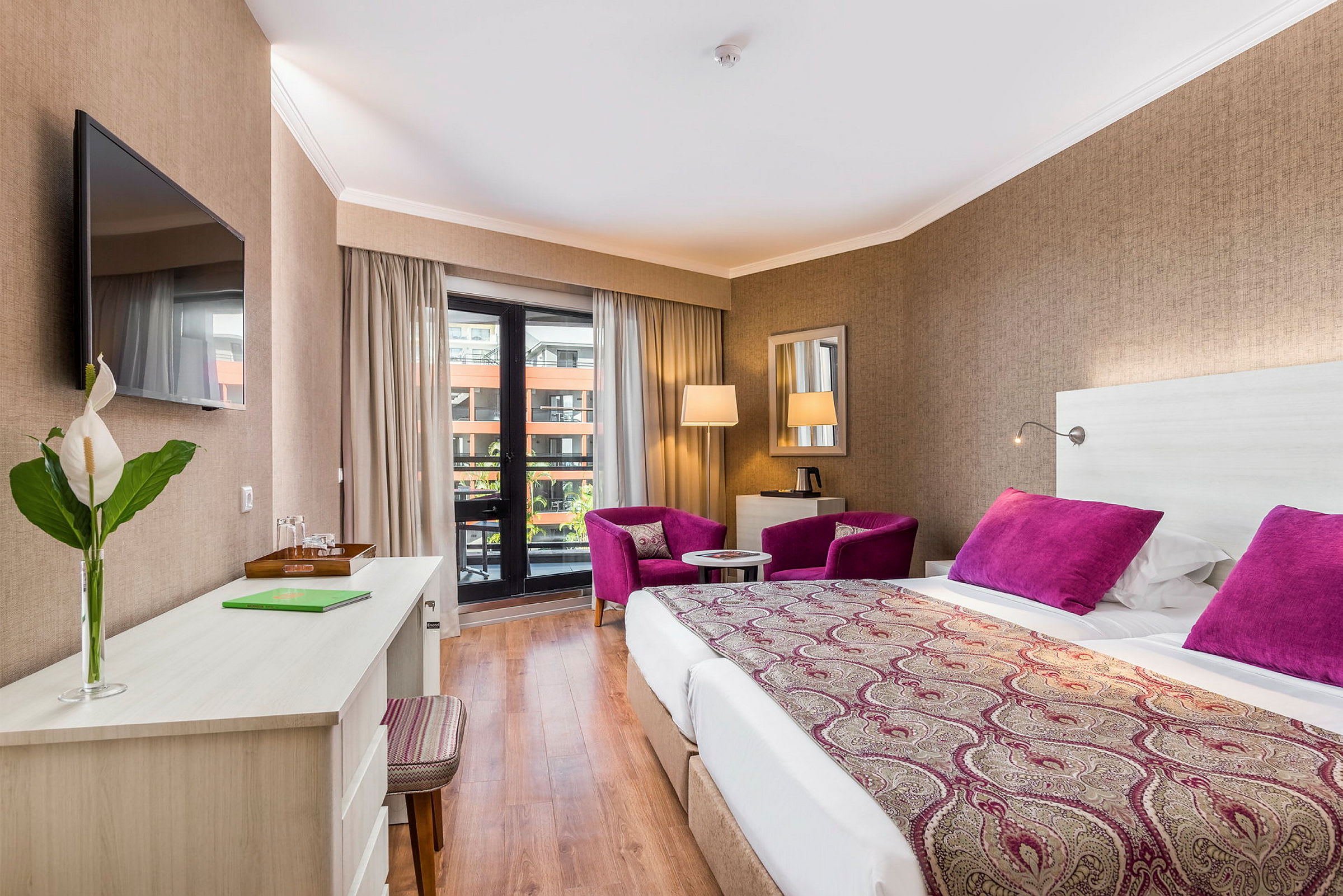 Hotellrom med dobbeltseng, skrivebord, to lilla lenestoler og balkong. (AI-generert tekst)